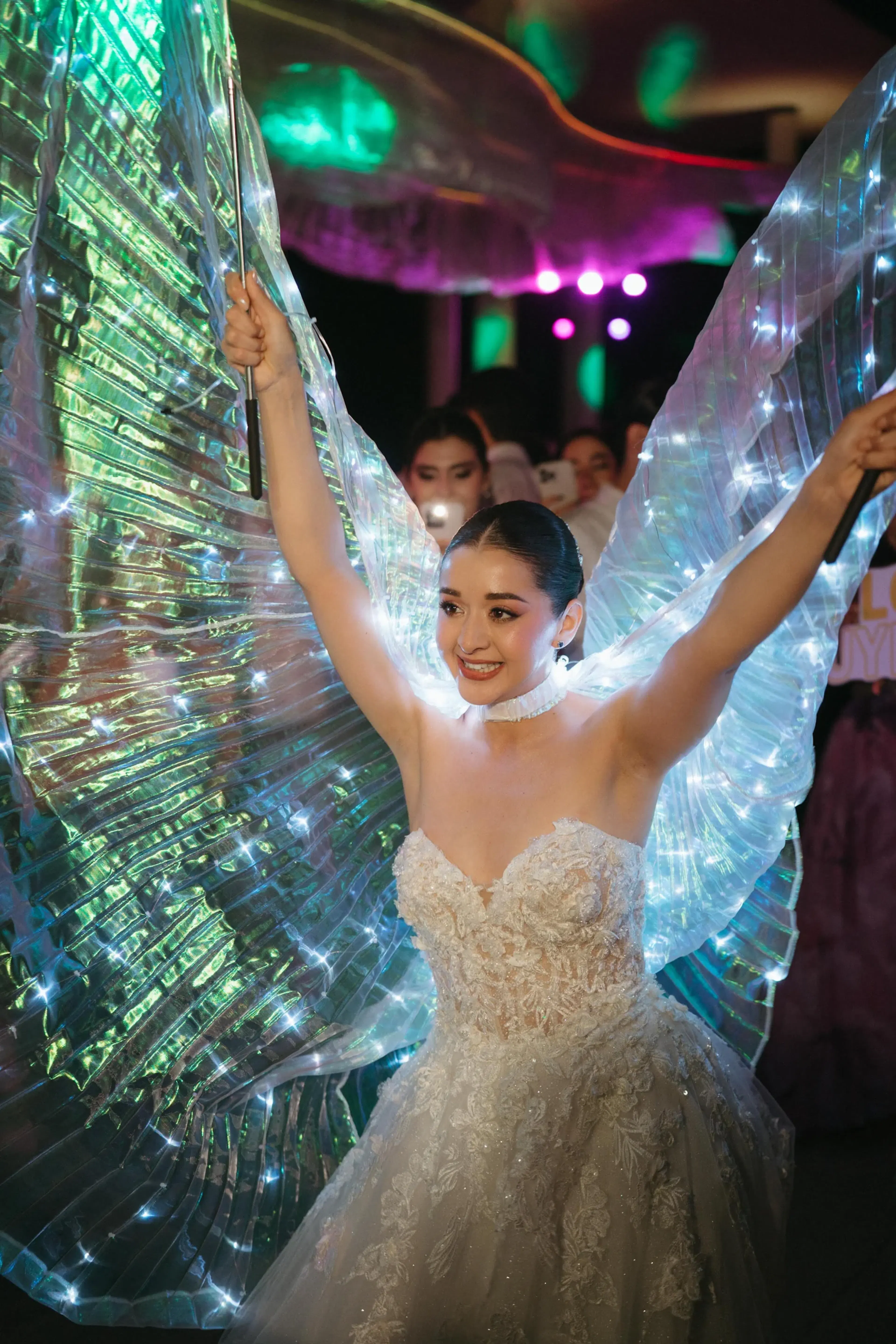 Una mujer con un vestido de encaje brillante y sin tirantes y alas iluminadas baila alegremente bajo luces de colores, transmitiendo una atmósfera festiva y mágica.