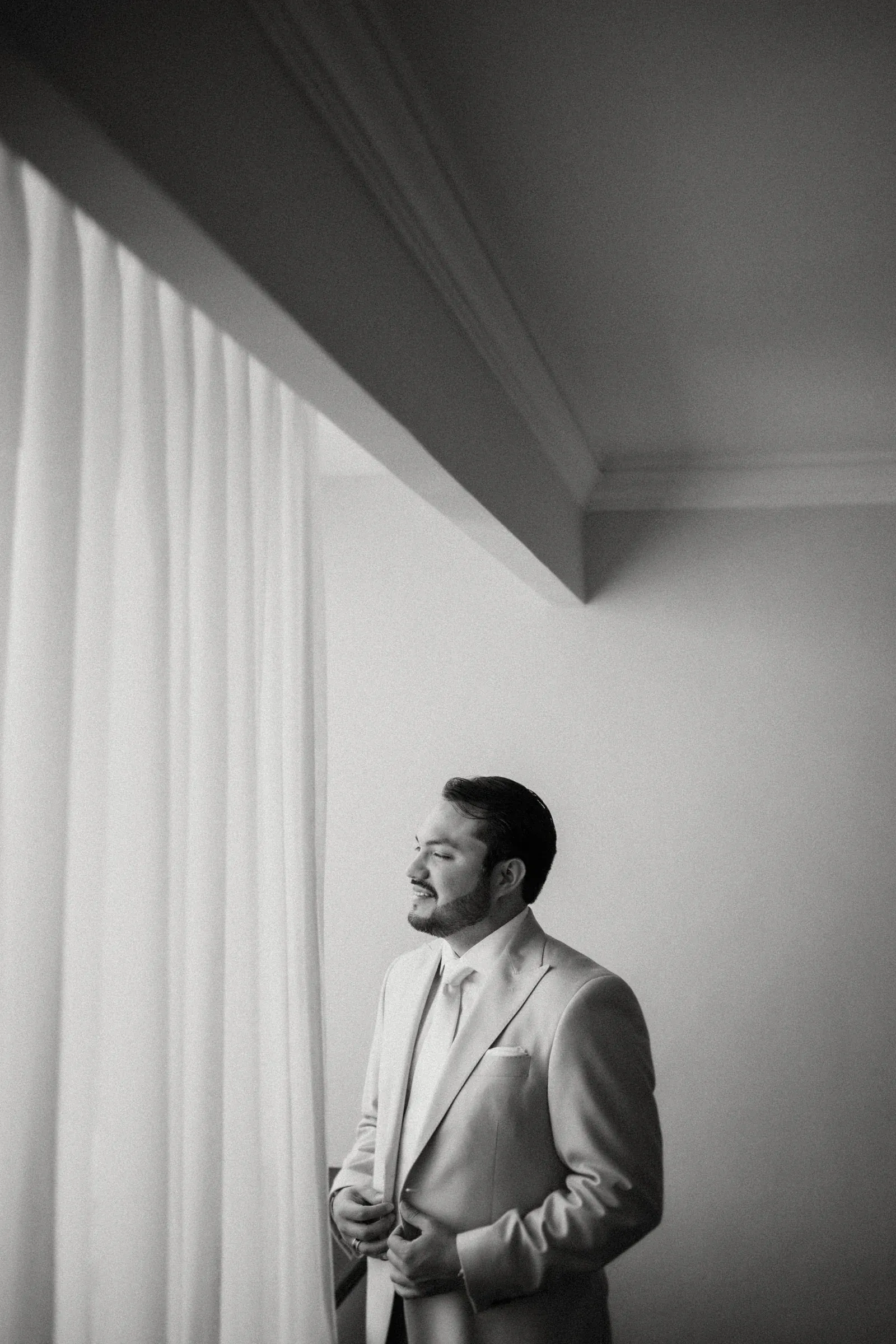 Un hombre de traje se encuentra junto a una ventana, abotonándose la chaqueta. La luz del sol se filtra suavemente a través de las cortinas, proyectando una atmósfera serena y contemplativa en blanco y negro.
