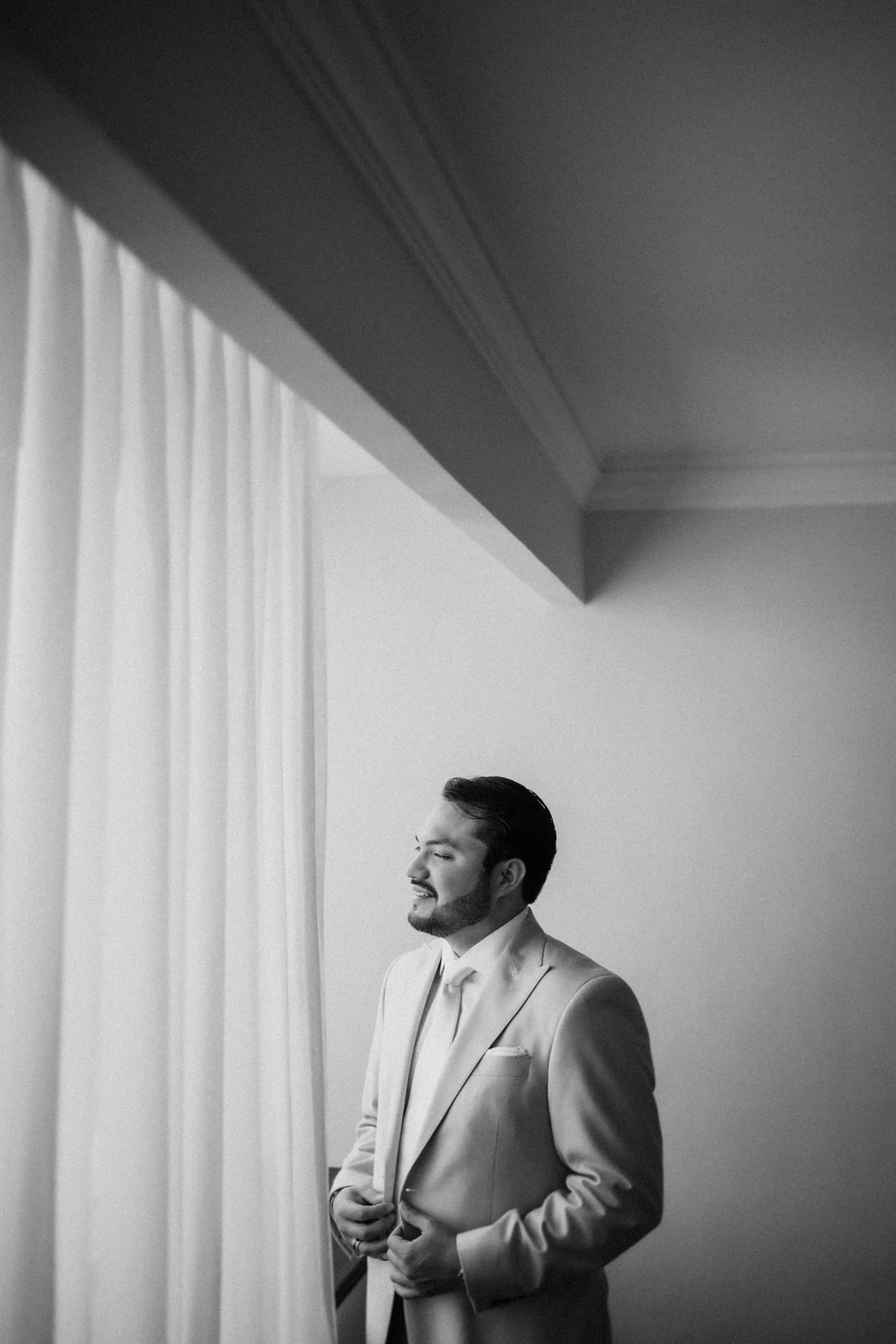 Un hombre de traje se encuentra junto a una ventana, abotonándose la chaqueta. La luz del sol se filtra suavemente a través de las cortinas, proyectando una atmósfera serena y contemplativa en blanco y negro.