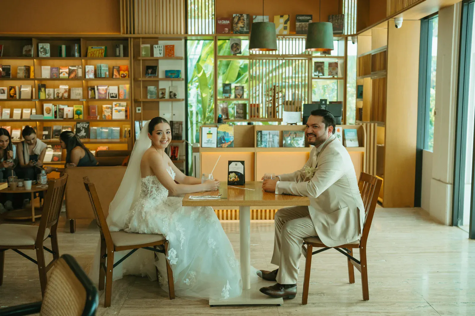 Los novios en una acogedora cafetería, sentados uno frente al otro en una mesa de madera. Estanterías llenas de libros y una iluminación tenue crean un ambiente cálido y romántico.