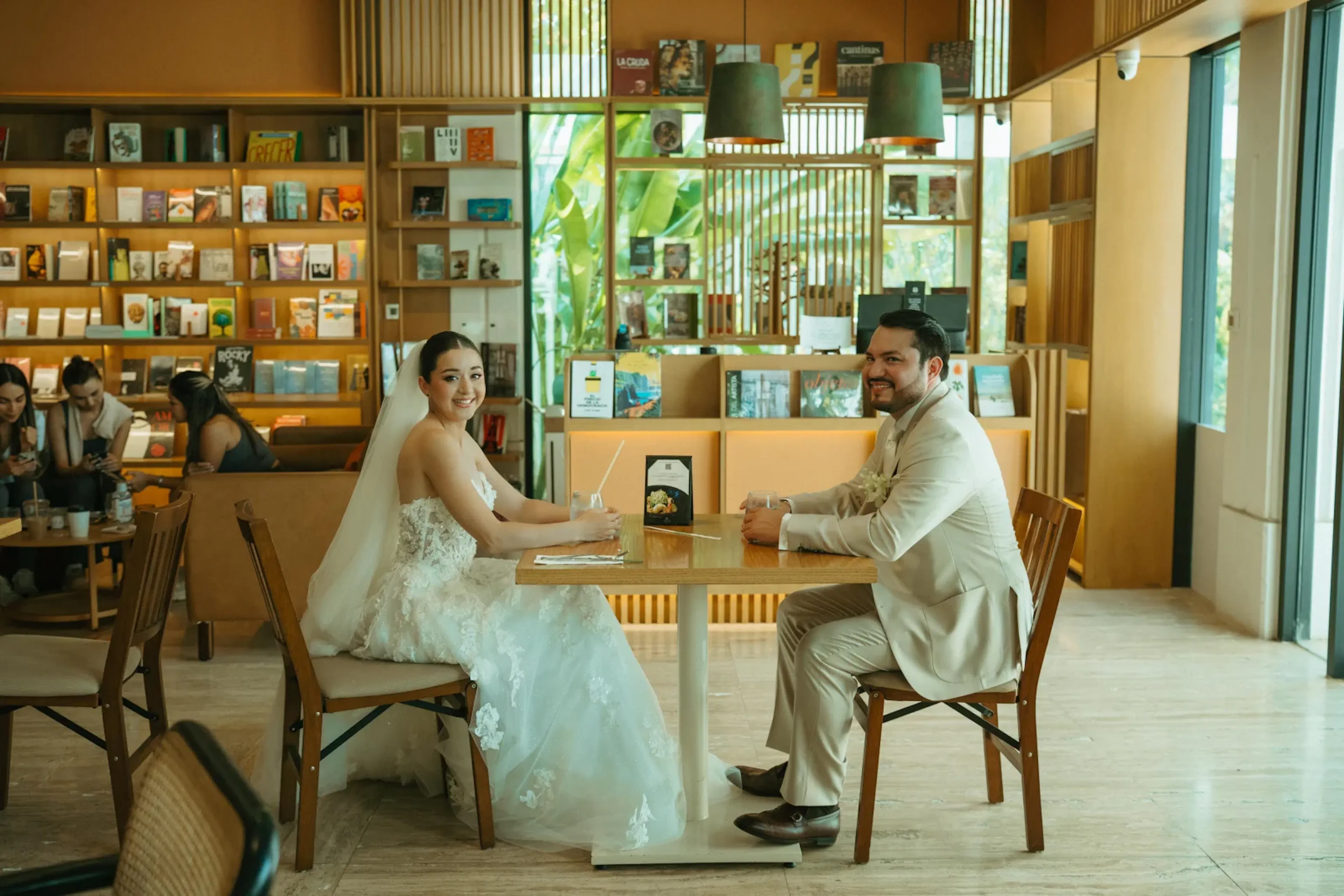 Los novios en una acogedora cafetería, sentados uno frente al otro en una mesa de madera. Estanterías llenas de libros y una iluminación tenue crean un ambiente cálido y romántico.