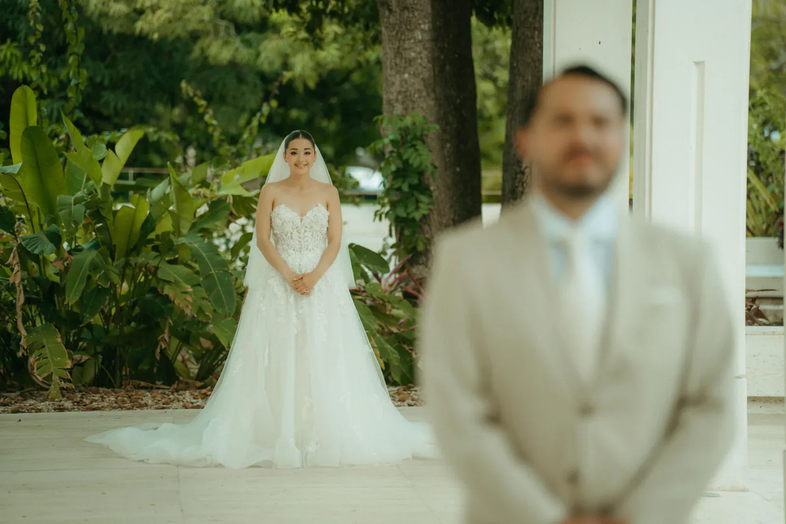 Una novia con un vestido de encaje blanco se yergue sonriendo en medio de una exuberante vegetación, mientras que un novio con un traje beige aparece borroso en primer plano, transmitiendo anticipación.