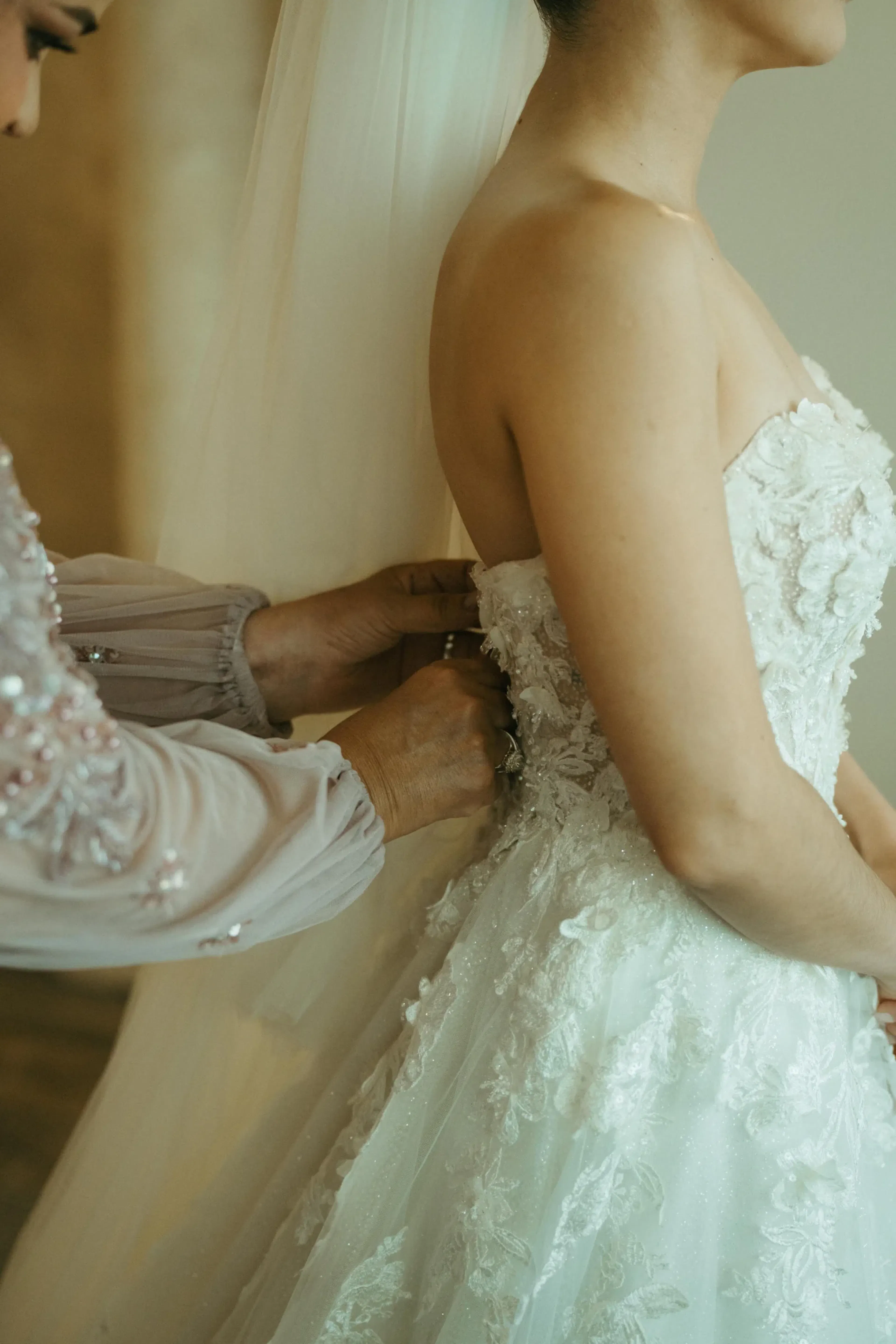 Una novia con un elegante vestido de novia de encaje blanco recibe ayuda con la parte de atrás de su vestido por una persona que viste un traje de manga larga con cuentas de color malva, lo que transmite una sensación de tierna preparación.