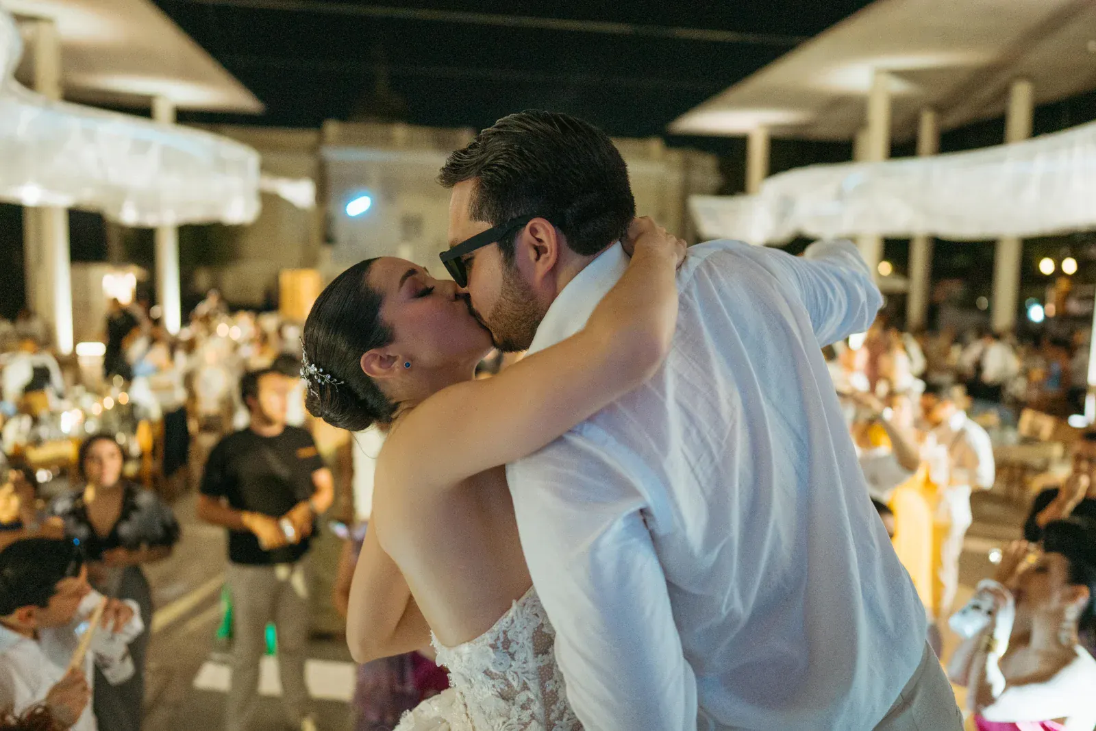 Los novios se besan en la pista de baile durante una boda al aire libre. Los invitados los rodean, celebrando bajo una suave iluminación ambiental.