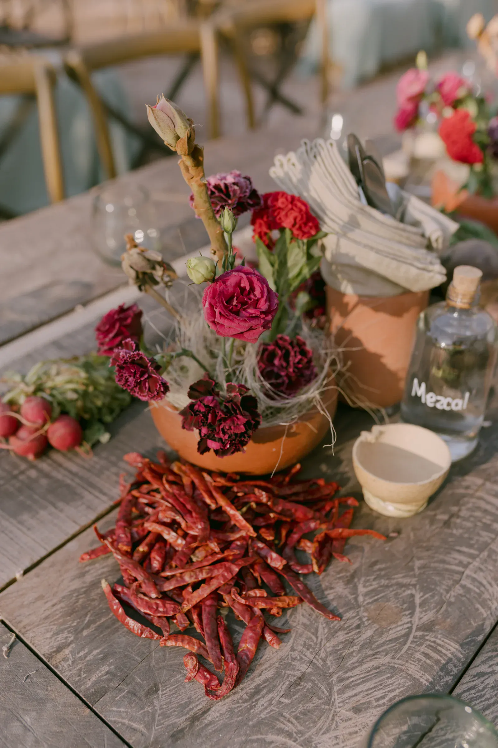 Una mesa rústica presenta una maceta de barro con vibrantes flores rojas y moradas, chiles rojos, rábanos, una botella de mezcal y servilletas dobladas, evocando calidez y festividad.