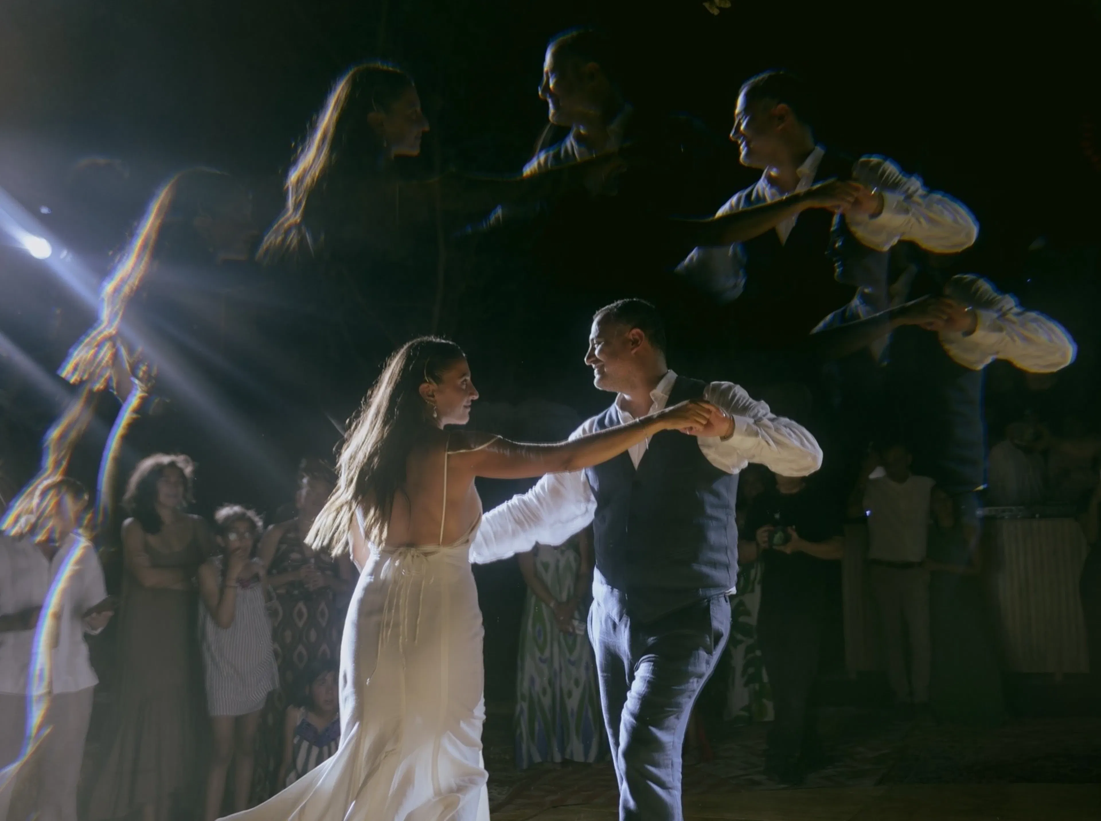 Una pareja de novios baila alegremente, bajo una suave luz. Los reflejos los rodean, con los invitados observándolos al fondo. Un ambiente romántico.