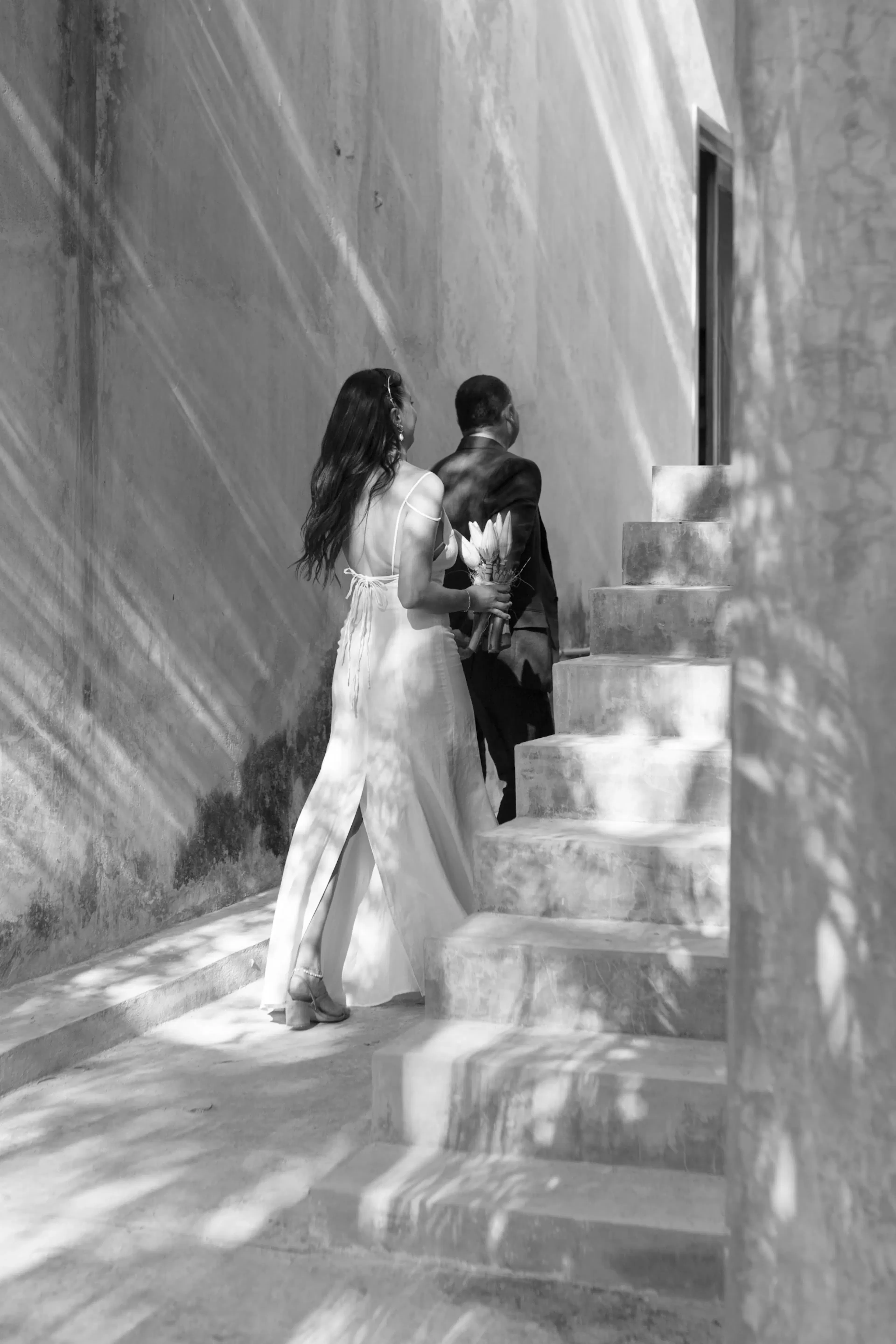 Una pareja subiendo unas escaleras de piedra. La mujer, con un vestido blanco vaporoso, sostiene un ramo de flores. La luz del sol, moteada, crea patrones en la pared. Tono romántico.