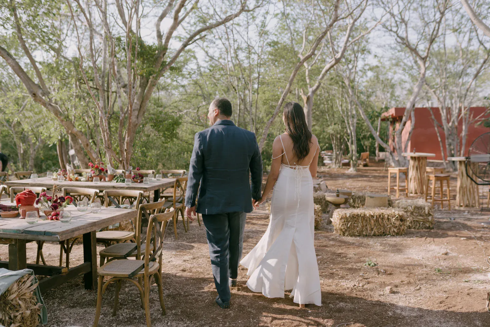 Una pareja camina de la mano por un rústico salón de bodas al aire libre. La novia lleva un vestido blanco y el novio un traje azul. Las mesas están decoradas con flores y rodeadas de árboles, creando un ambiente sereno y romántico.