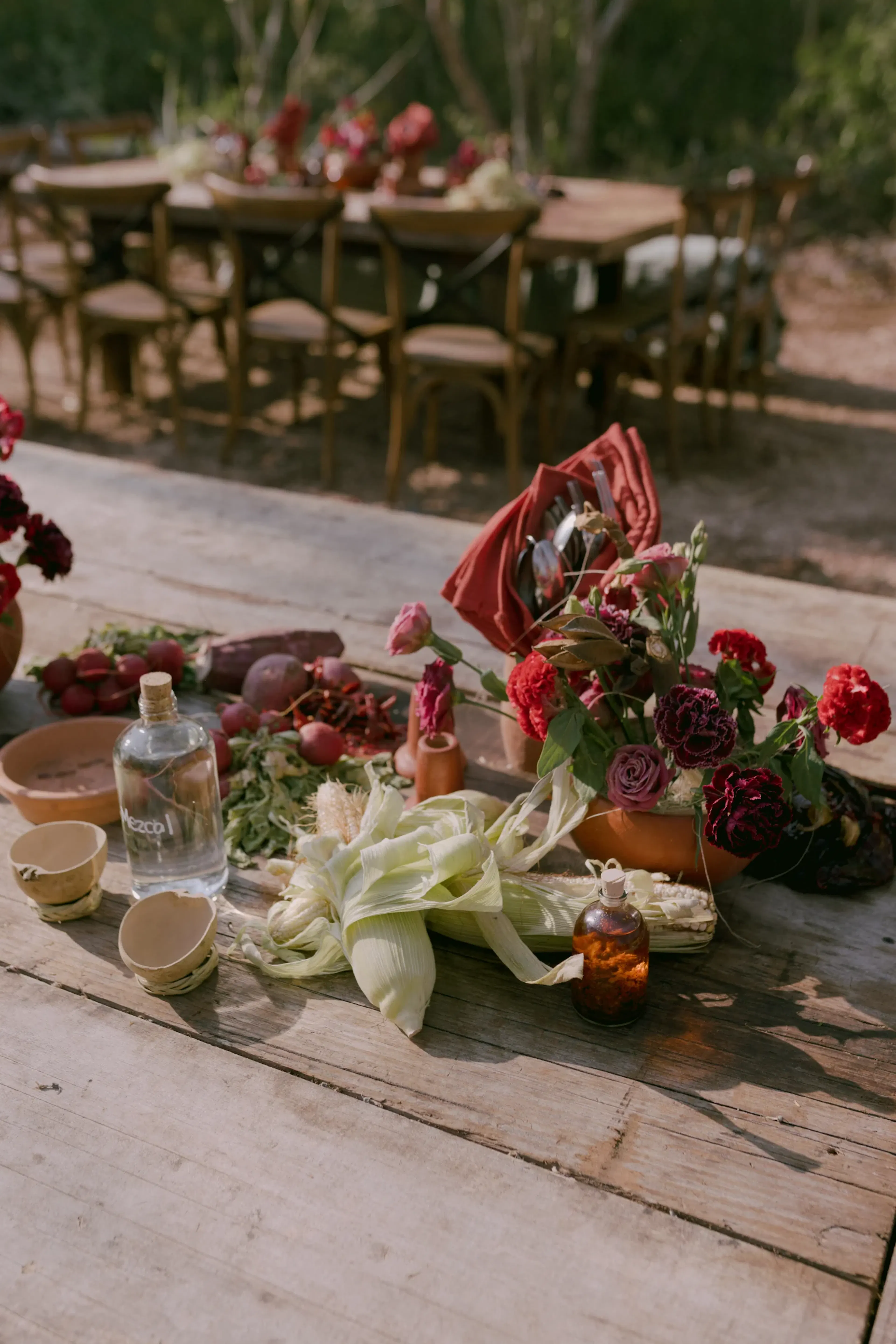 Una mesa rústica de exterior está adornada con vibrantes flores rojas, maíz fresco, una botella de agua y pequeños cuencos. Un comedor más amplio con sillas de madera se difumina al fondo, creando un ambiente cálido y natural.
