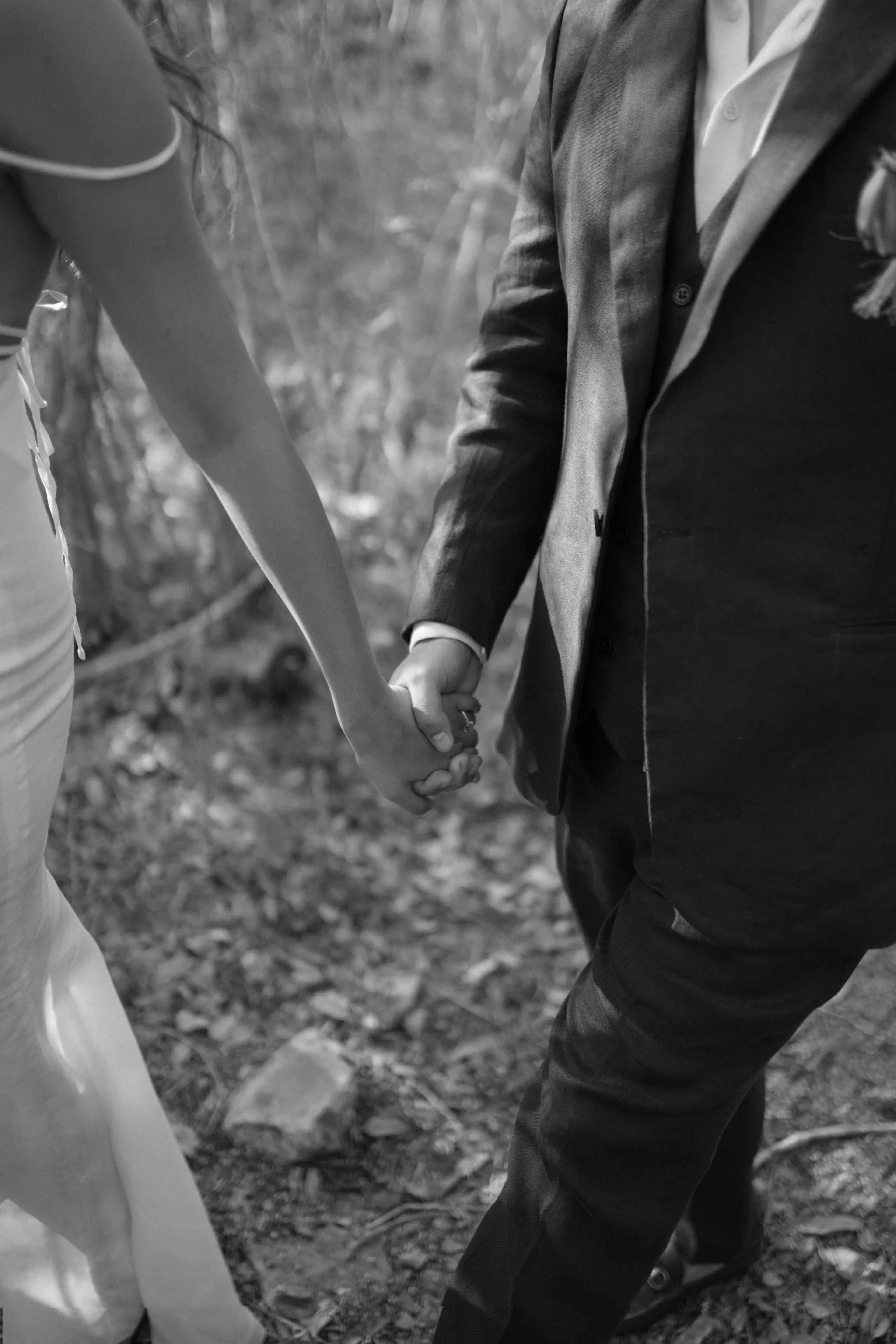 Una pareja de novios, tomados de la mano, camina por un sendero forestal. La novia lleva un vestido blanco y el novio un traje oscuro. Una romántica e íntima foto en blanco y negro.