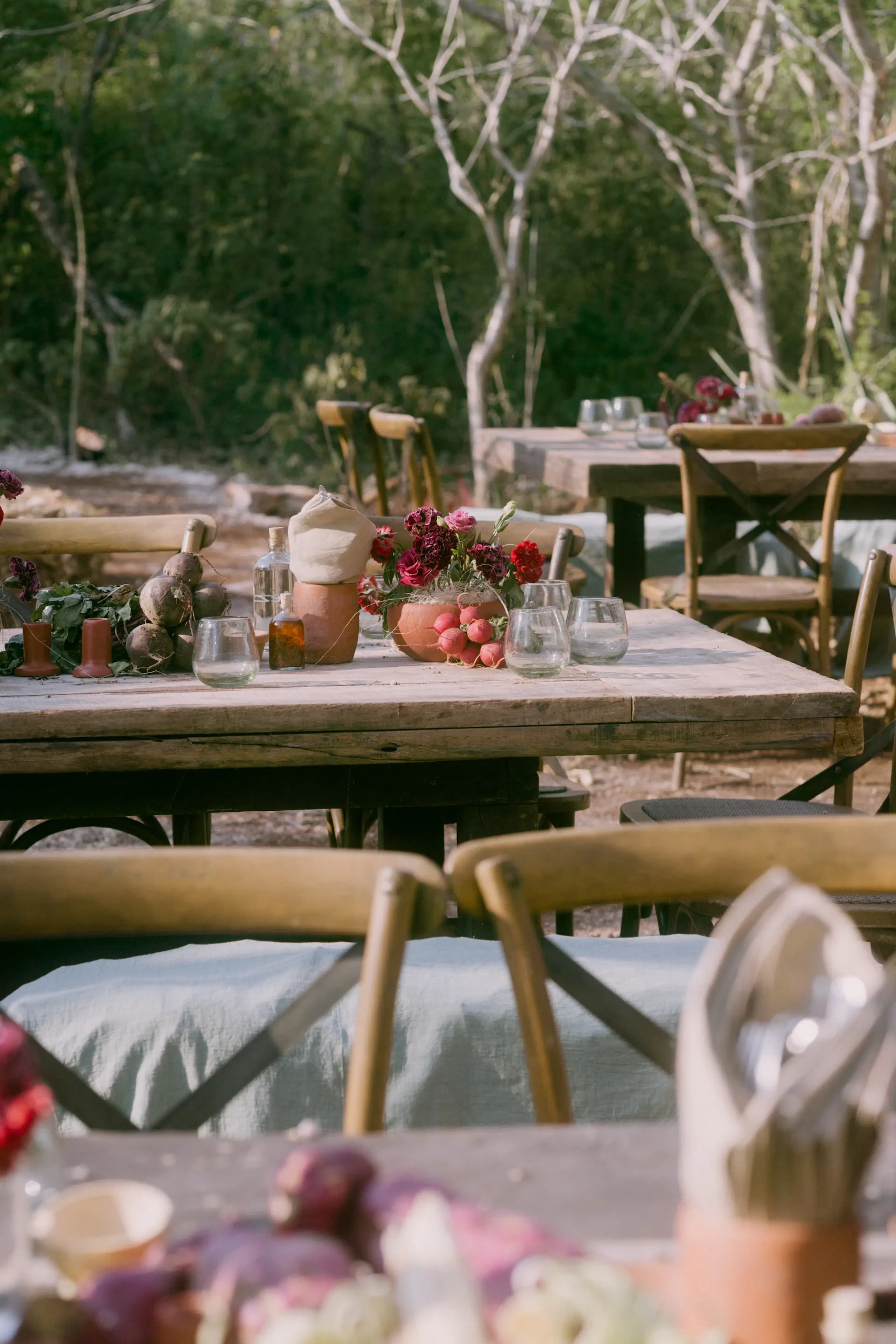 Comedor rústico al aire libre con mesas y sillas de madera, adornado con vibrantes flores rojas y rosas, velas y cristalería transparente, rodeado de árboles.