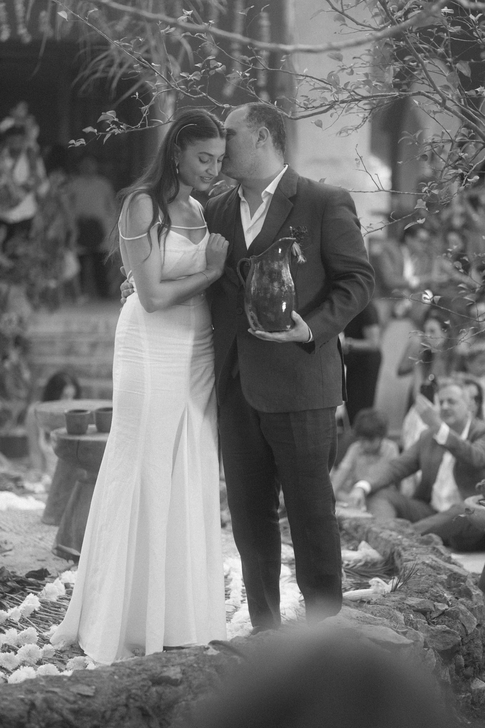Una pareja con atuendo formal comparte un momento íntimo en una ceremonia. El hombre besa la frente de la mujer mientras sostiene una jarra decorativa. Una atmósfera serena y amorosa los rodea, con los invitados borrosos al fondo.