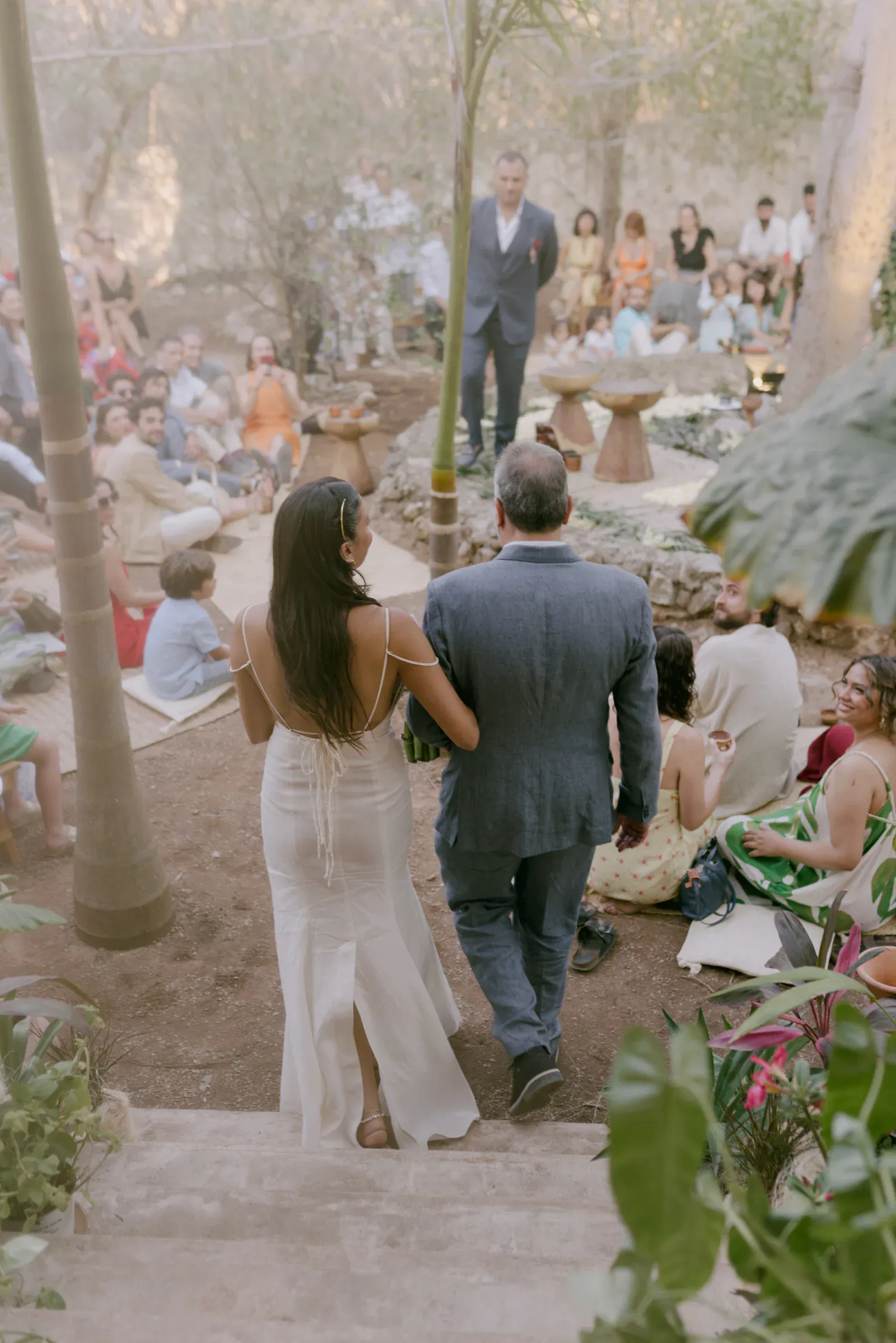 Una novia con vestido blanco baja las escaleras del brazo de un hombre en una boda al aire libre. Los invitados se sientan tranquilamente en el suelo, creando un ambiente cálido e íntimo.