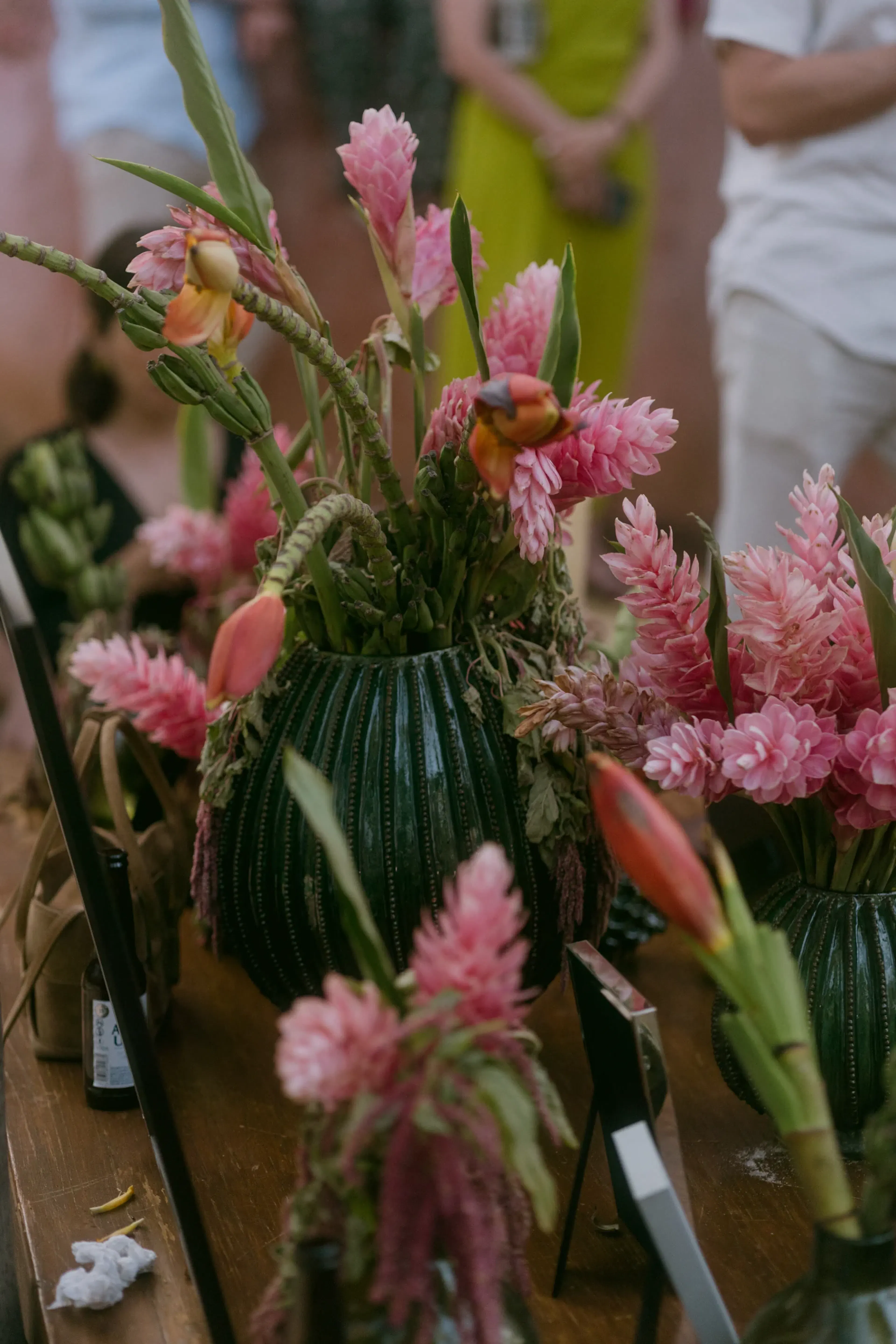 Jarrones de cerámica verde con vibrantes flores rosas y naranjas sobre una mesa de madera. Al fondo se ven figuras borrosas con trajes coloridos.