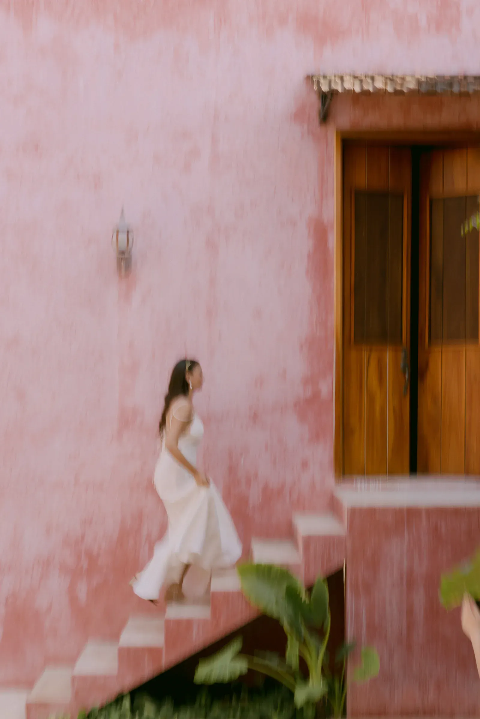Una mujer con un vaporoso vestido blanco sube una escalera de estuco rosa. Al fondo, una puerta rústica de madera y vegetación evocan un ambiente tranquilo y tropical.