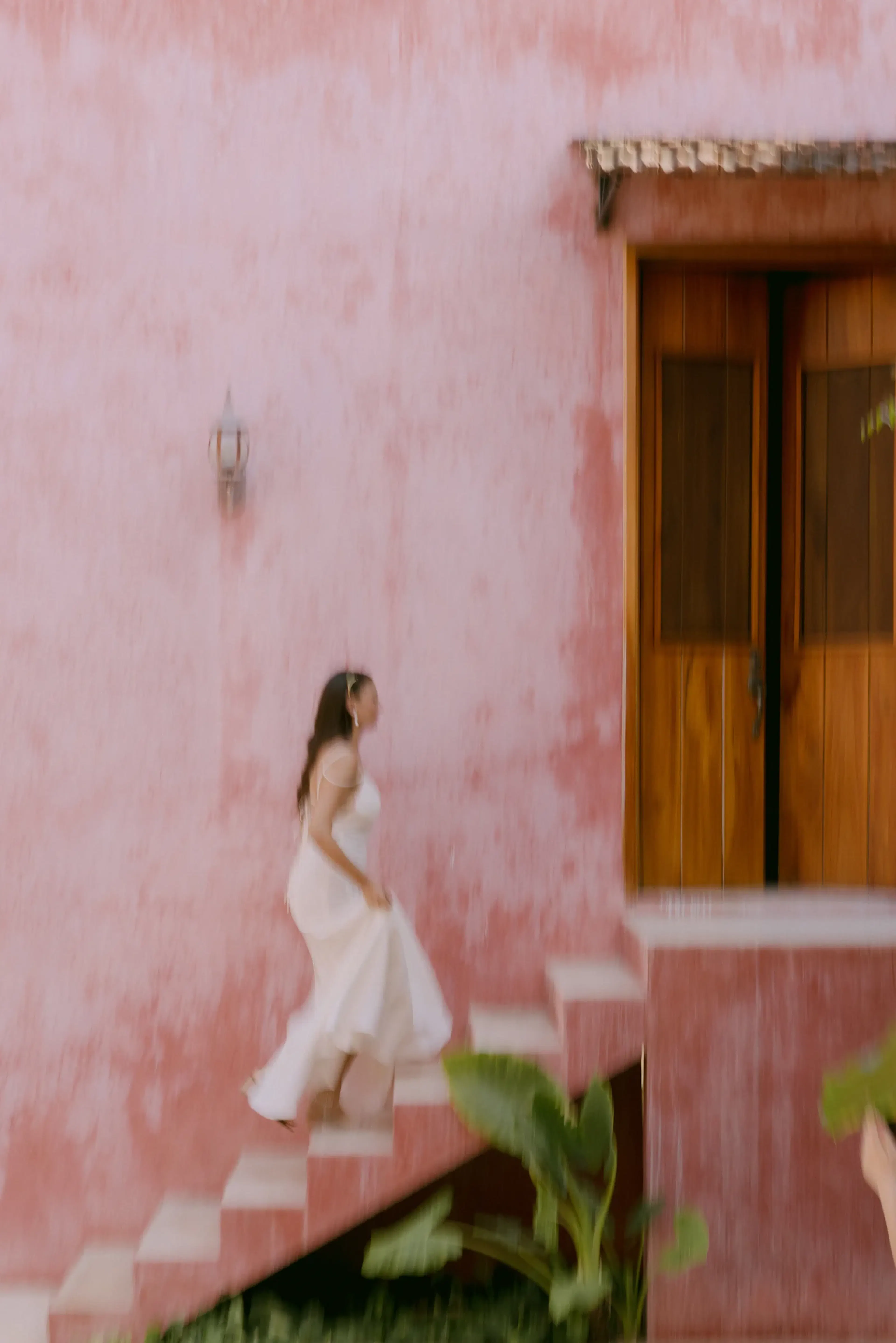 Una mujer con un vaporoso vestido blanco sube una escalera de estuco rosa. Al fondo, una puerta rústica de madera y vegetación evocan un ambiente tranquilo y tropical.