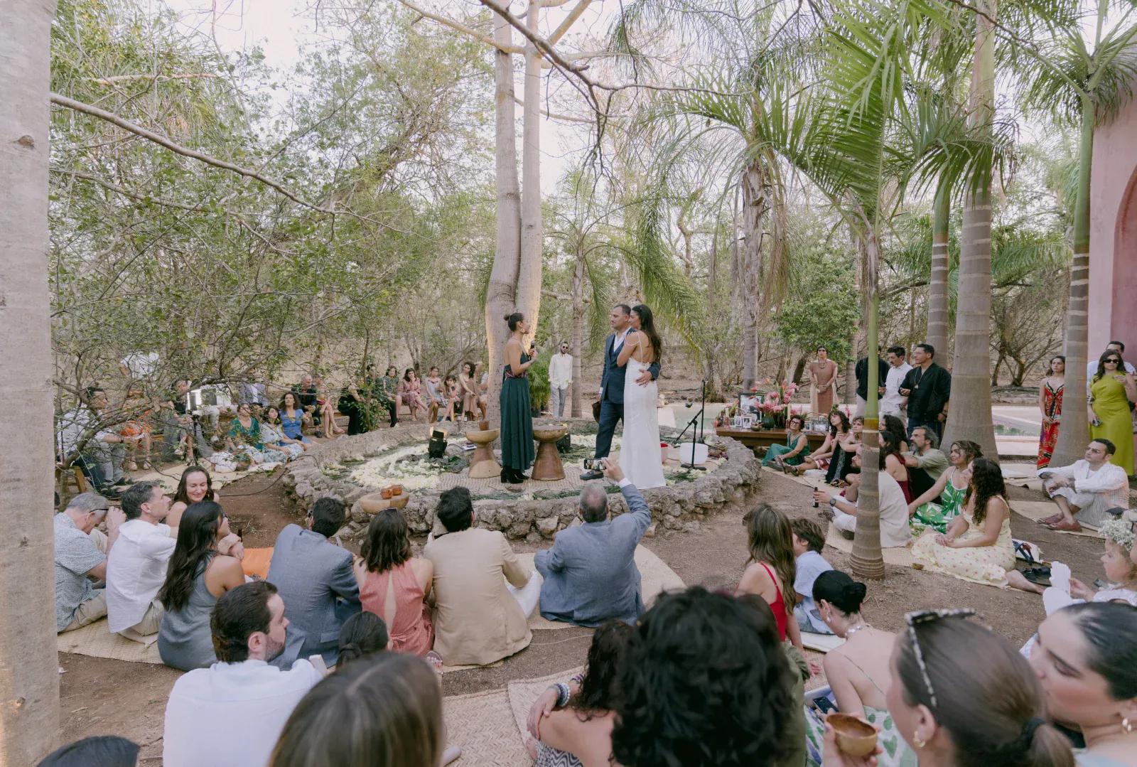 Una ceremonia de boda al aire libre en un bosque con una pareja abrazándose. Los invitados se sientan en el suelo, rodeándolos, creando un ambiente íntimo y alegre.