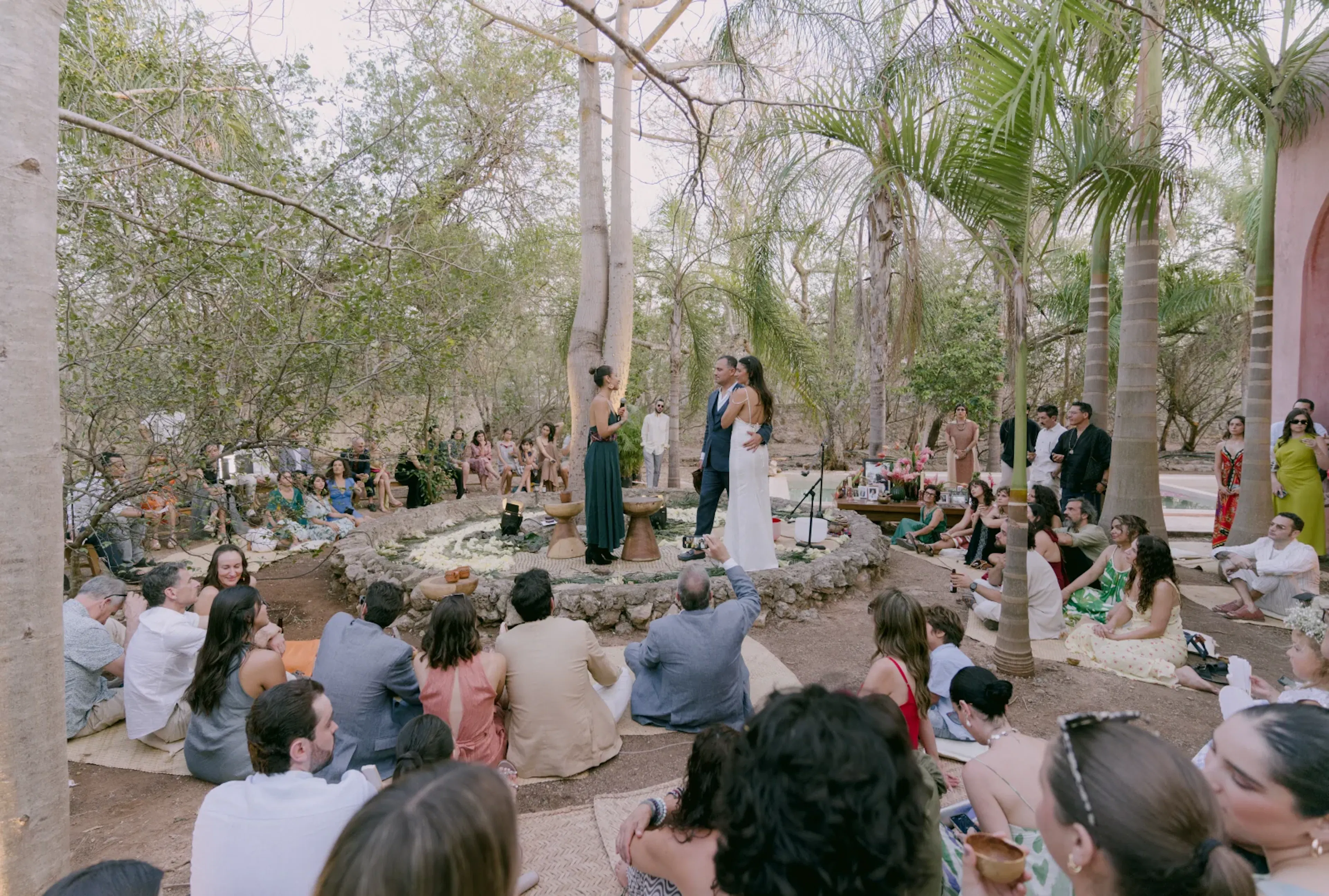 Una ceremonia de boda al aire libre en un bosque con una pareja abrazándose. Los invitados se sientan en el suelo, rodeándolos, creando un ambiente íntimo y alegre.
