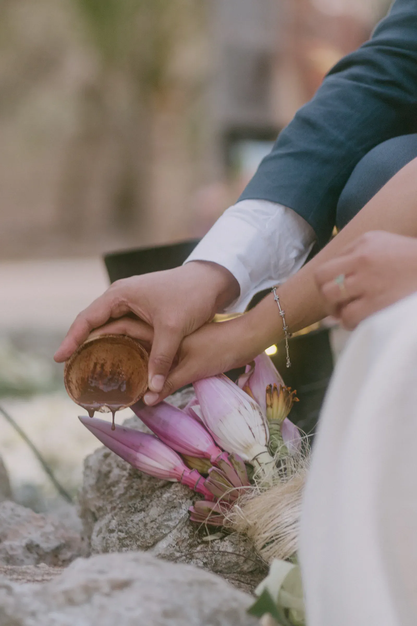 Primer plano de unas manos en un acto ceremonial, con agua vertida desde una taza de madera sobre flores de loto moradas, transmitiendo una sensación de tradición y serenidad.