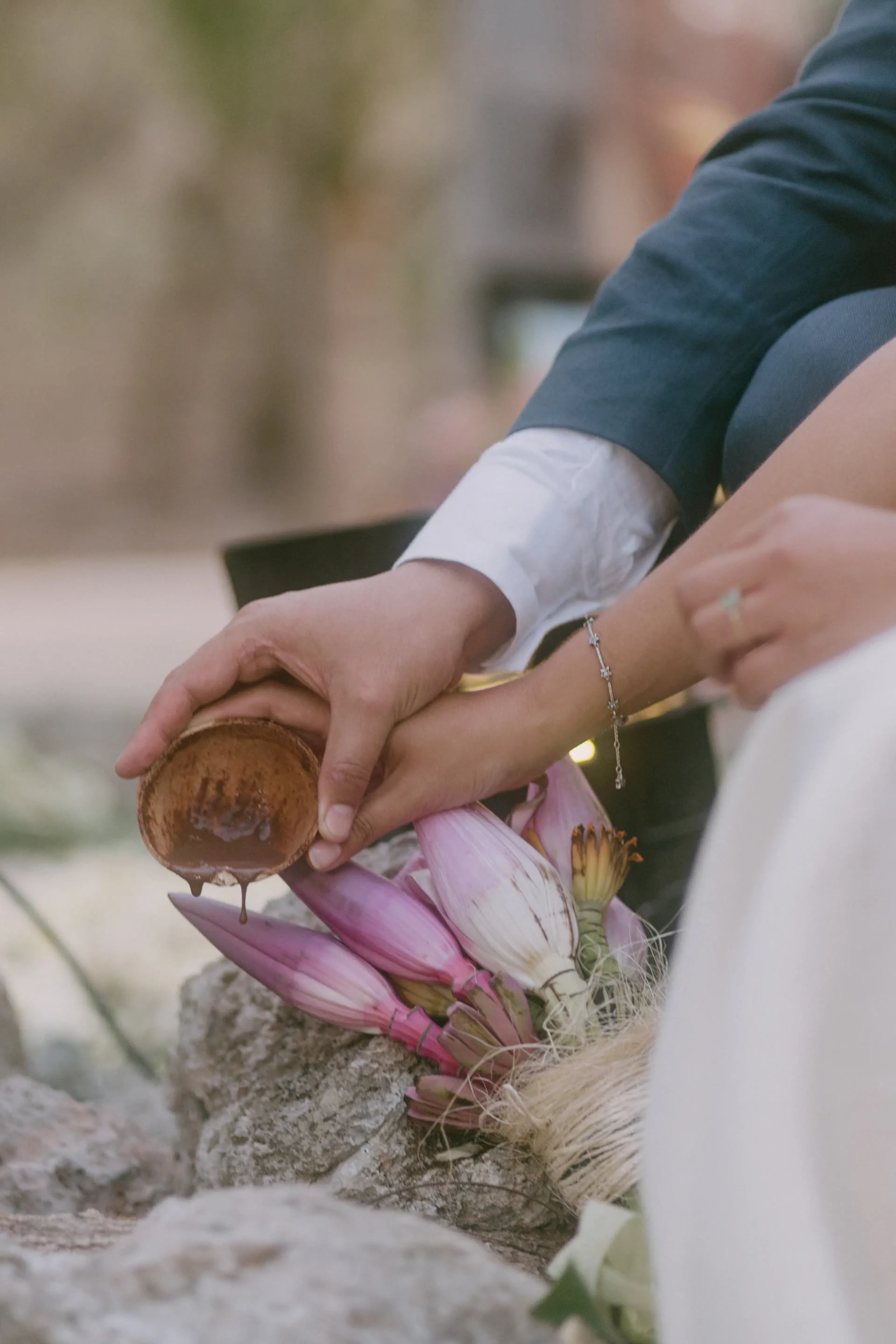 Primer plano de unas manos en un acto ceremonial, con agua vertida desde una taza de madera sobre flores de loto moradas, transmitiendo una sensación de tradición y serenidad.