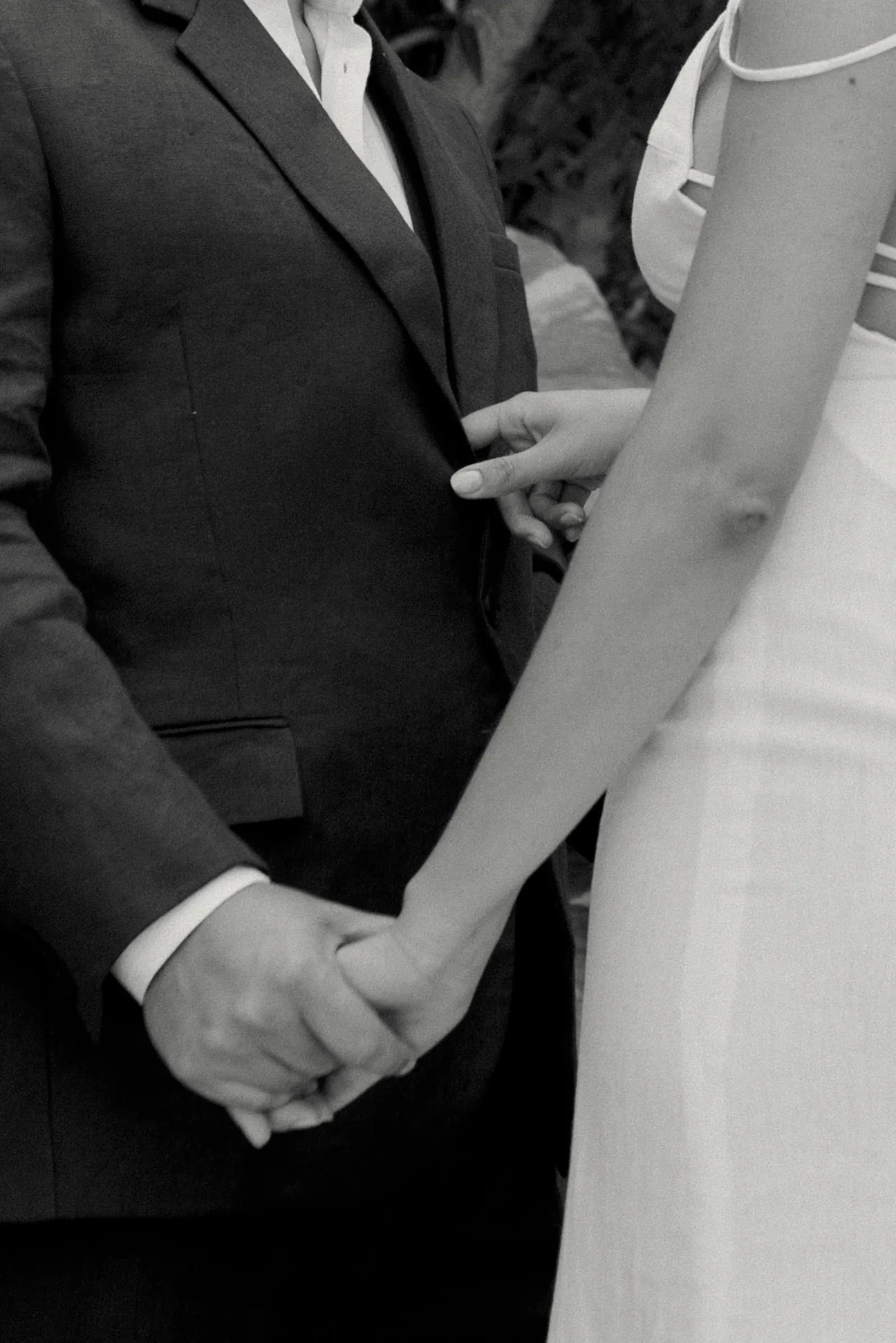 Una foto en blanco y negro muestra a una pareja tomada de la mano. La persona de la izquierda lleva traje y la de la derecha, vestido. La escena transmite intimidad y conexión.