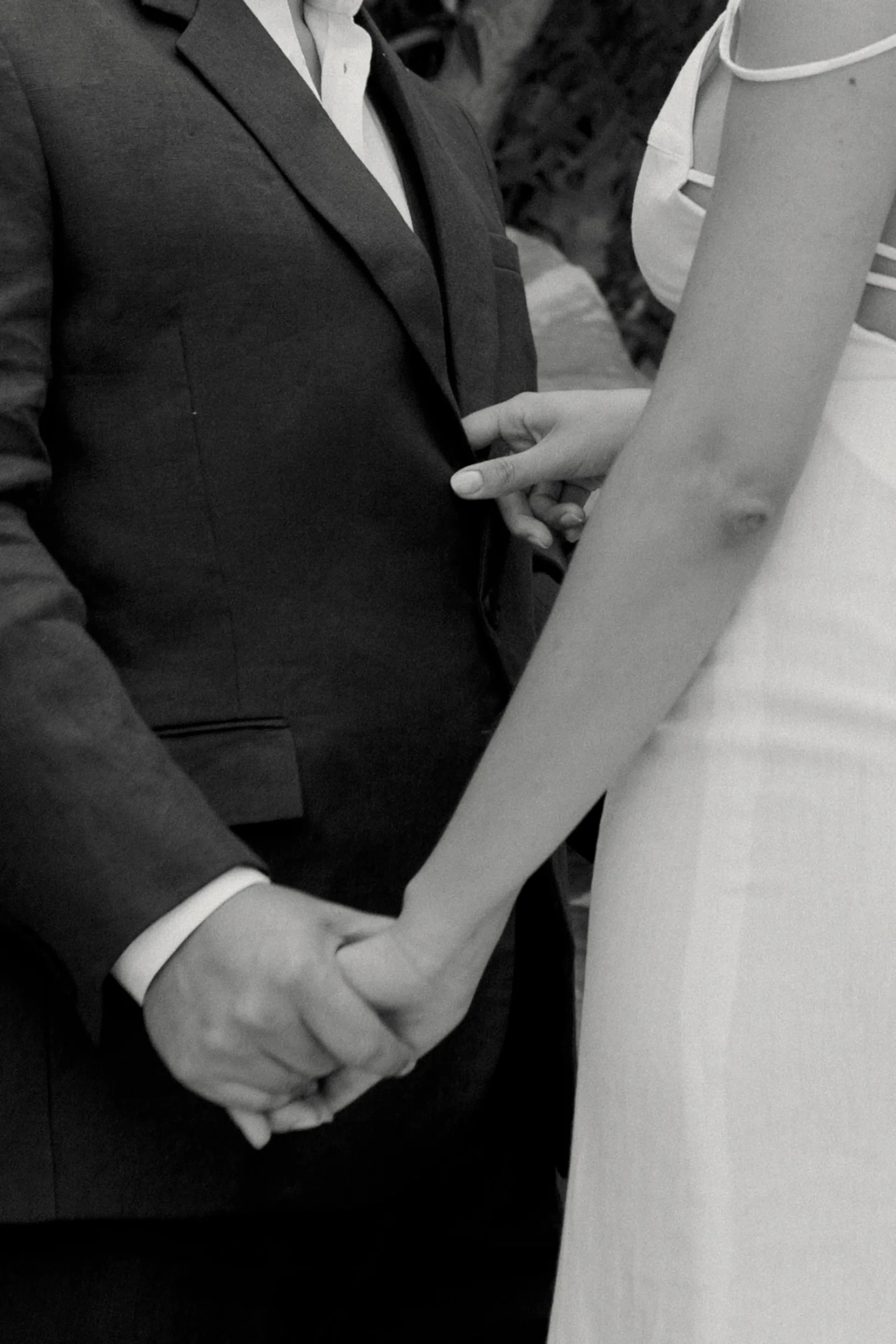 Una foto en blanco y negro muestra a una pareja tomada de la mano. La persona de la izquierda lleva traje y la de la derecha, vestido. La escena transmite intimidad y conexión.