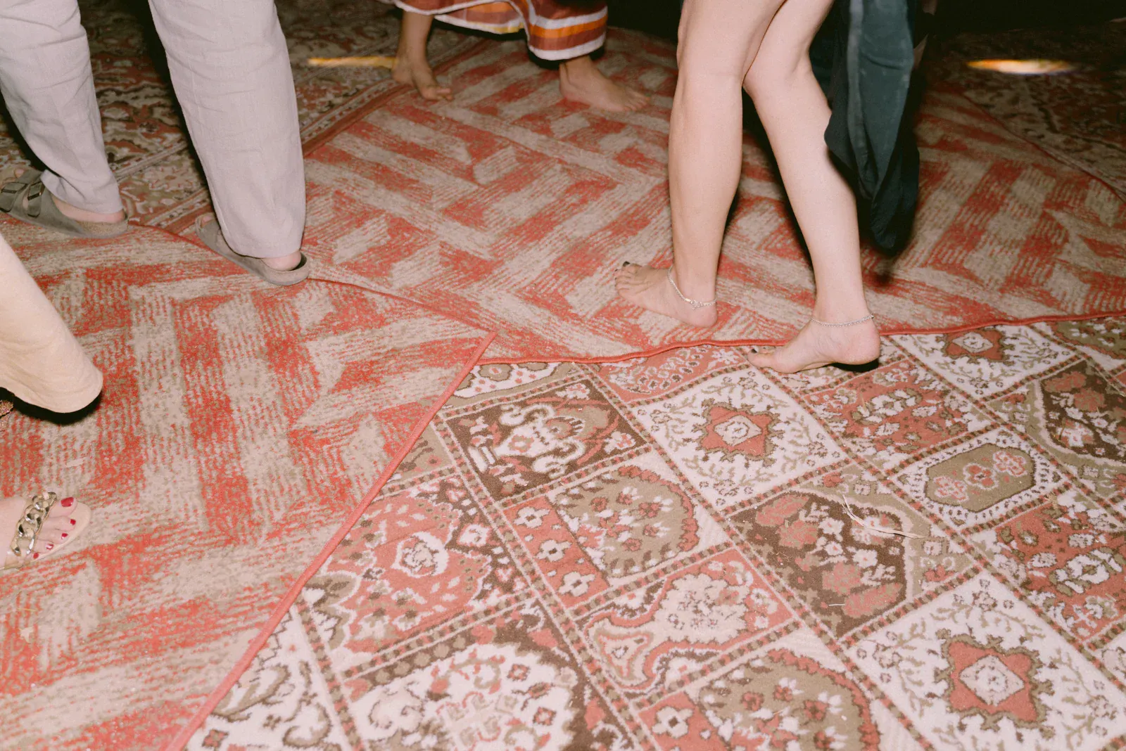 La gente baila descalza sobre alfombras con diseños geométricos y florales. La escena transmite una atmósfera relajada y alegre con una iluminación tenue.