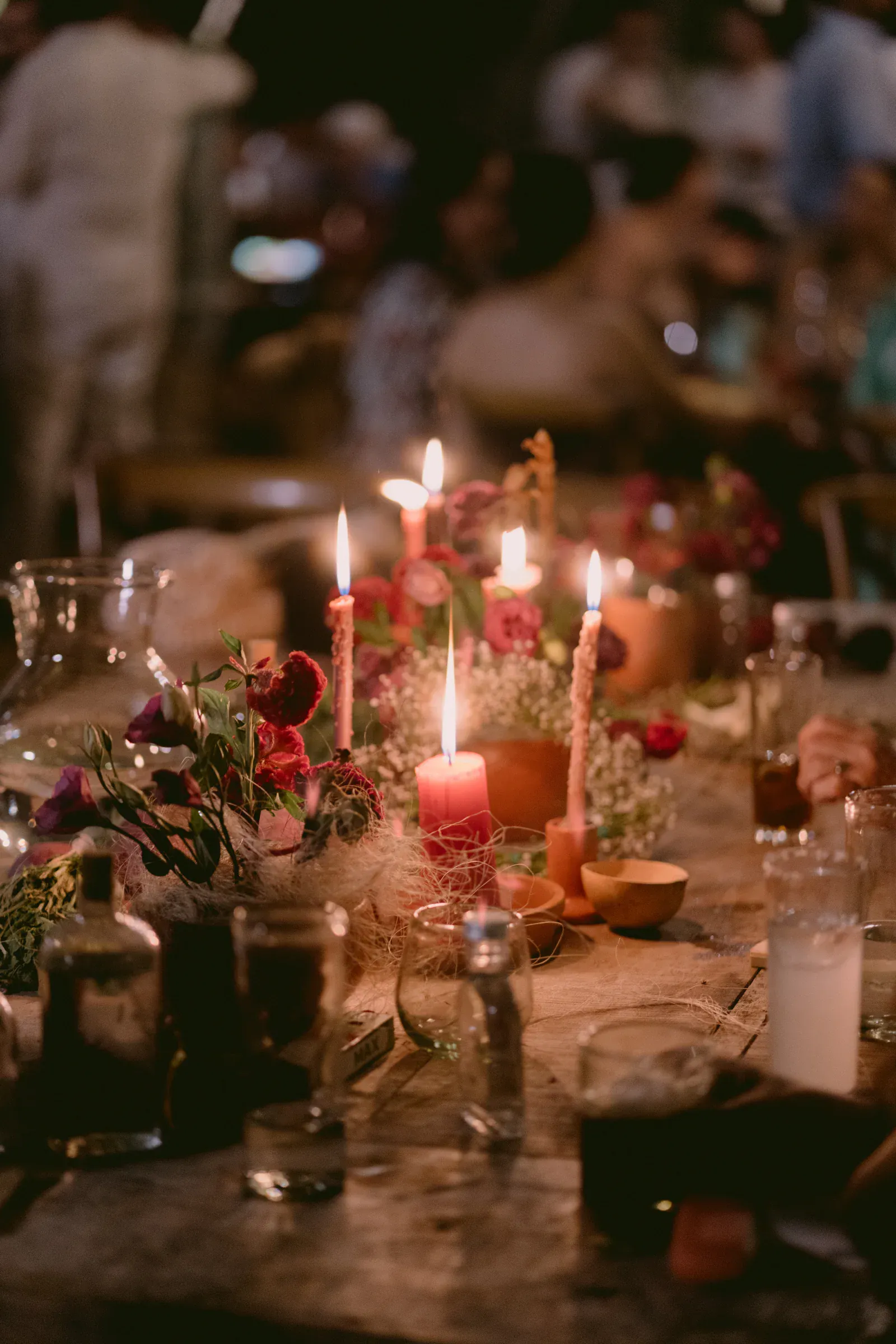 Una mesa rústica está adornada con velas brillantes, flores rojas y cristalería, creando un ambiente cálido en una reunión animada con invitados borrosos en el fondo.