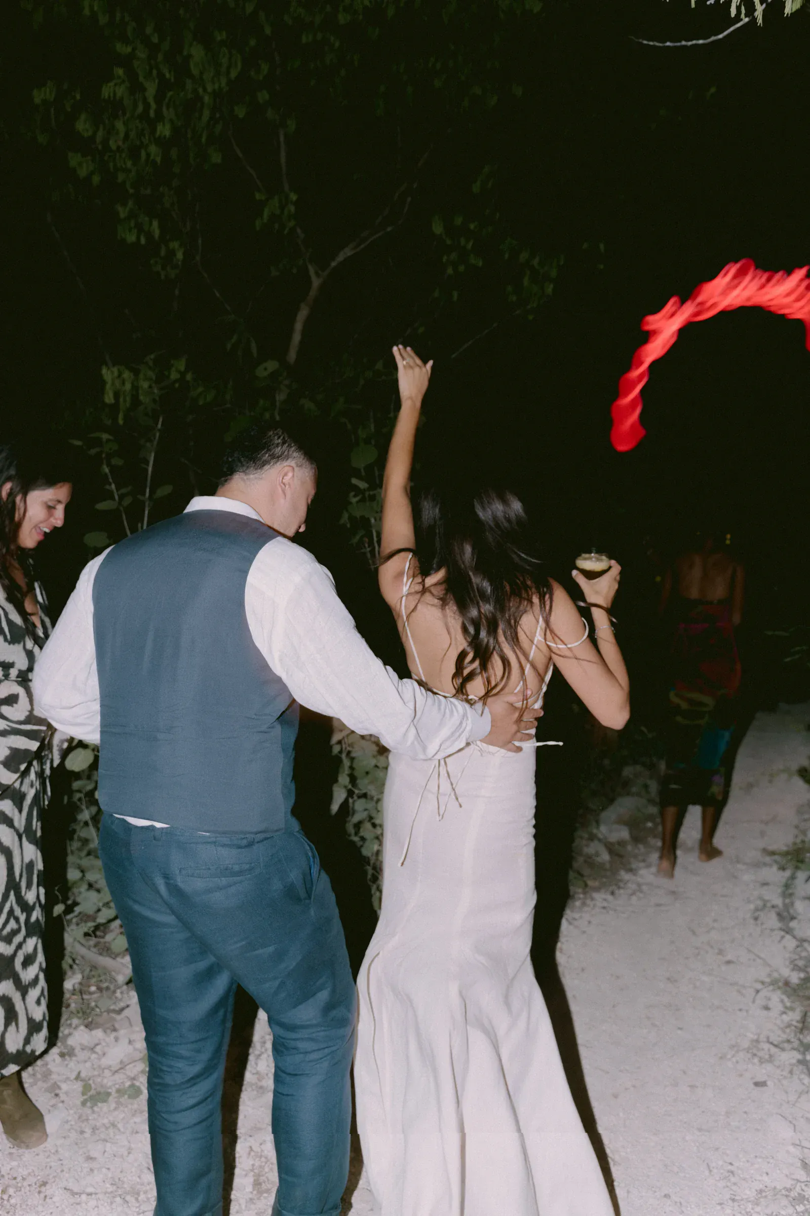Una pareja camina de la mano por un sendero tenuemente iluminado, rodeado de árboles. La mujer lleva un vestido con la espalda al aire y sostiene una bebida, evocando un ambiente alegre y festivo.