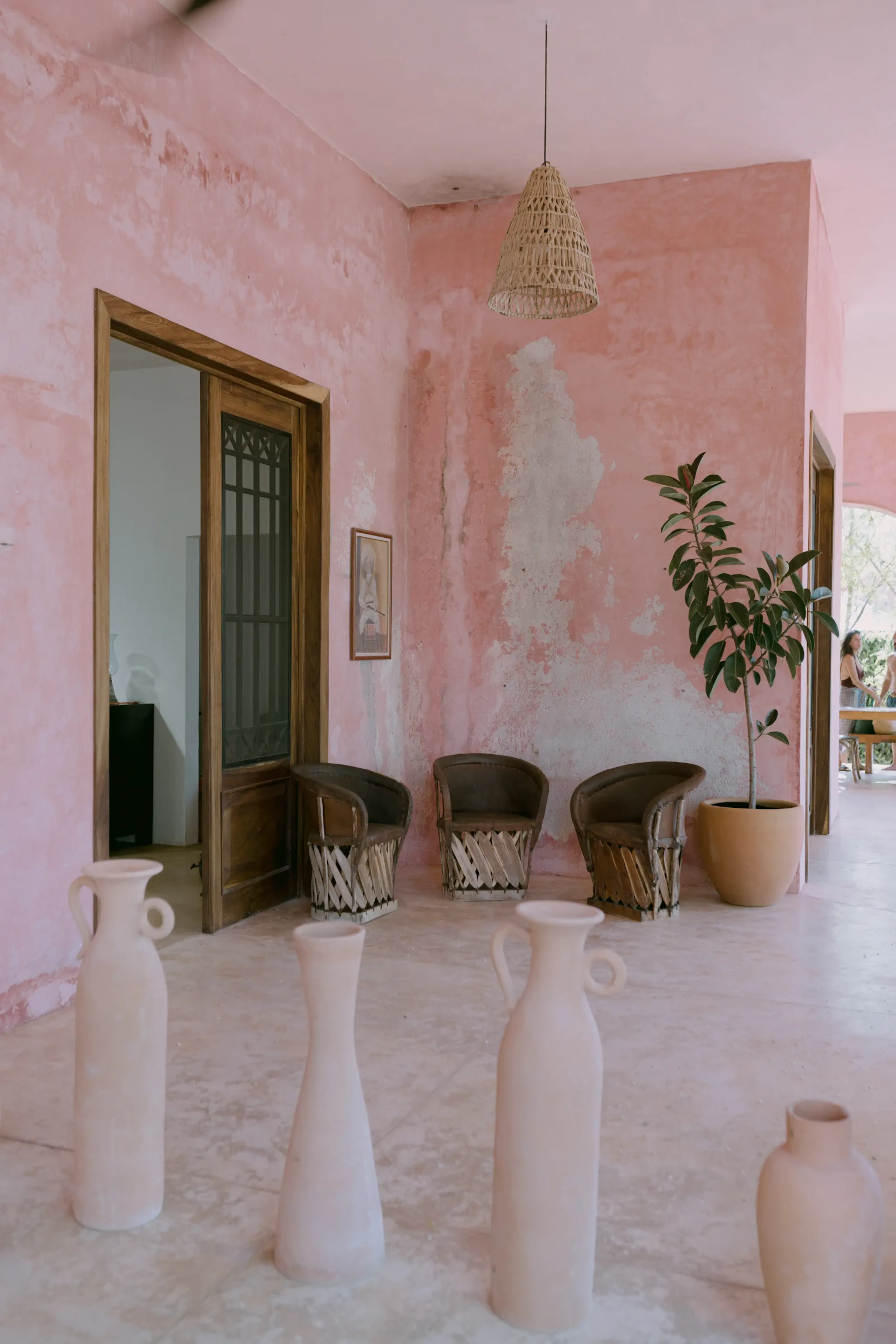 Habitación con paredes texturizadas rosas y sillas de madera. Una maceta y una lámpara tejida aportan calidez. Jarrones altos de cerámica adornan el primer plano. Ambiente relajado.
