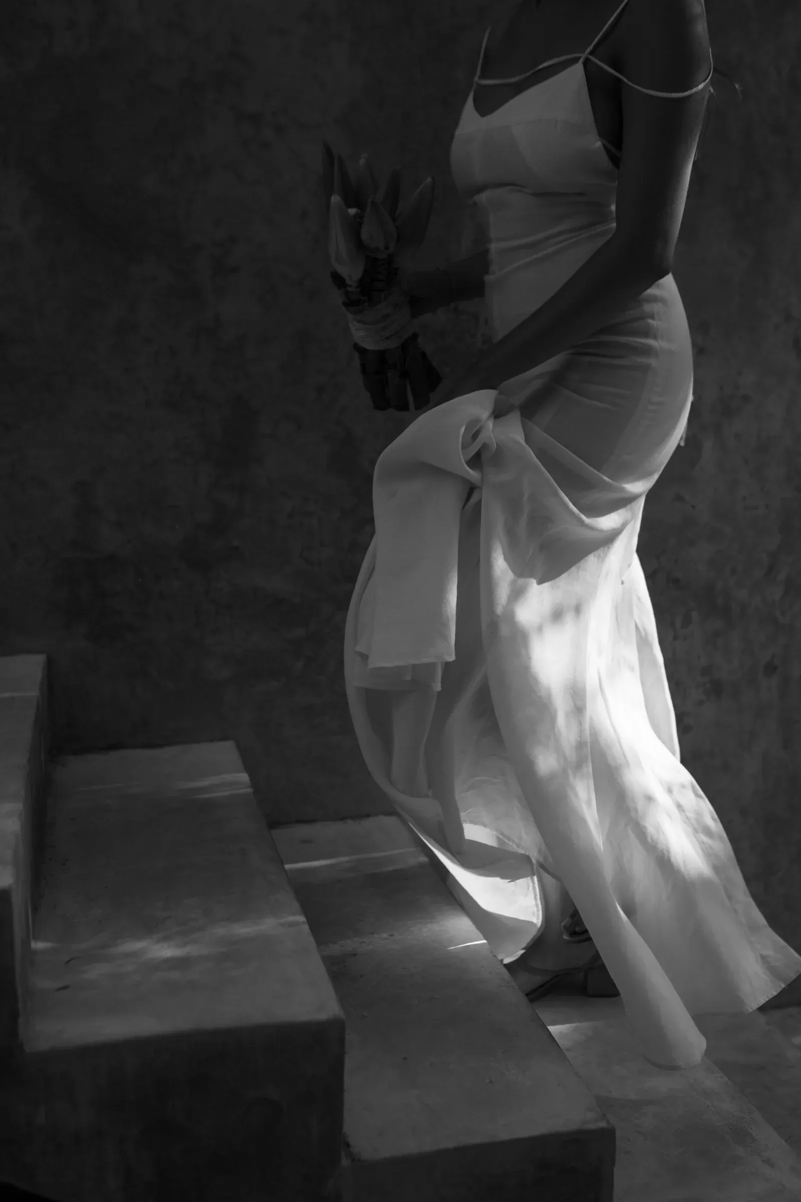 Una mujer con un vaporoso vestido blanco sube unas escaleras de piedra, sosteniendo un ramo de flores. Una luz tenue y moteada proyecta sombras sutiles, creando un ambiente sereno y elegante.