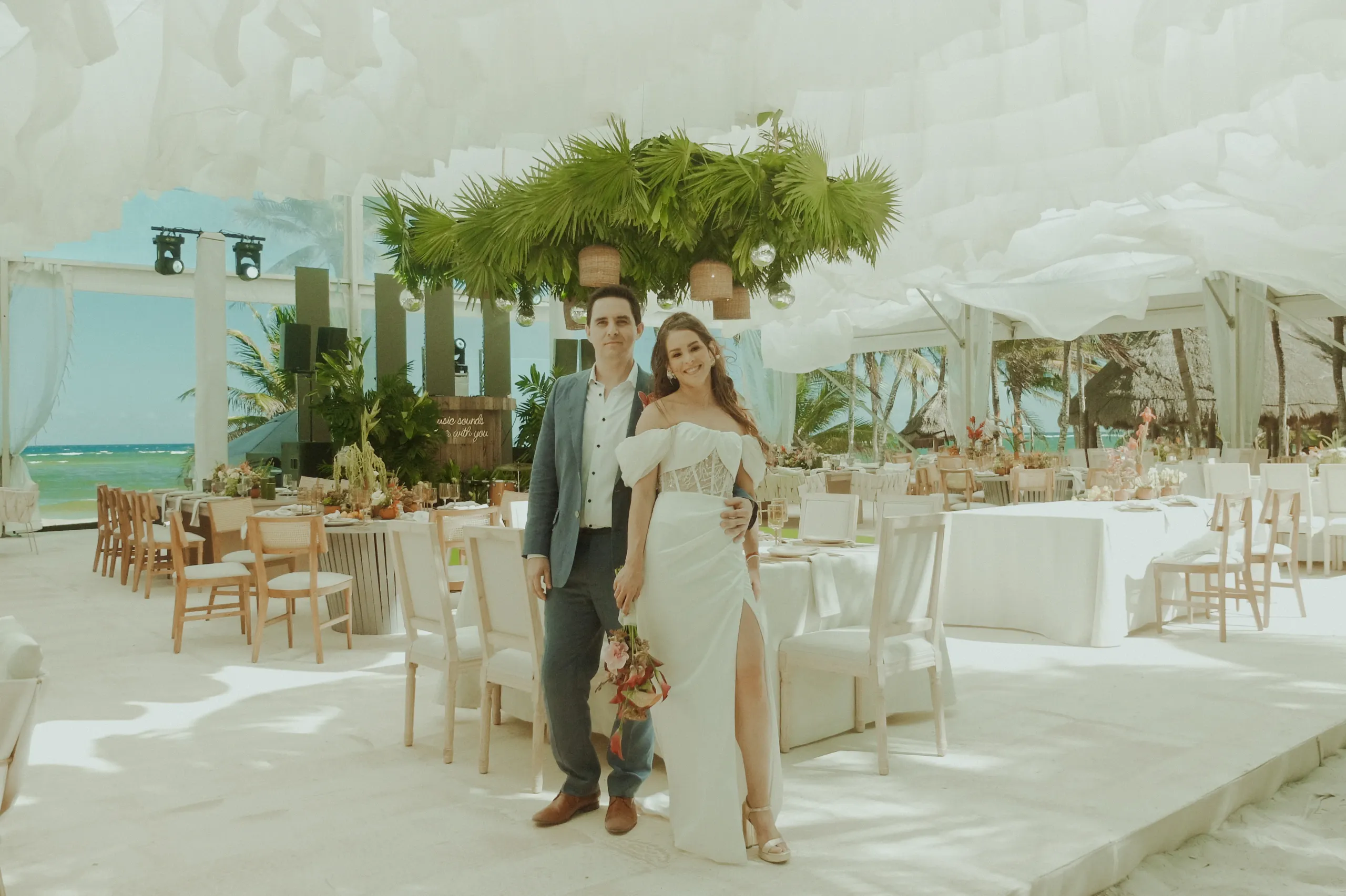 Una pareja de recién casados sonriendo, la novia con un vestido blanco y un ramo de flores, junto al novio con traje. Un lugar elegante y espacioso con vistas al mar y decoración tropical.