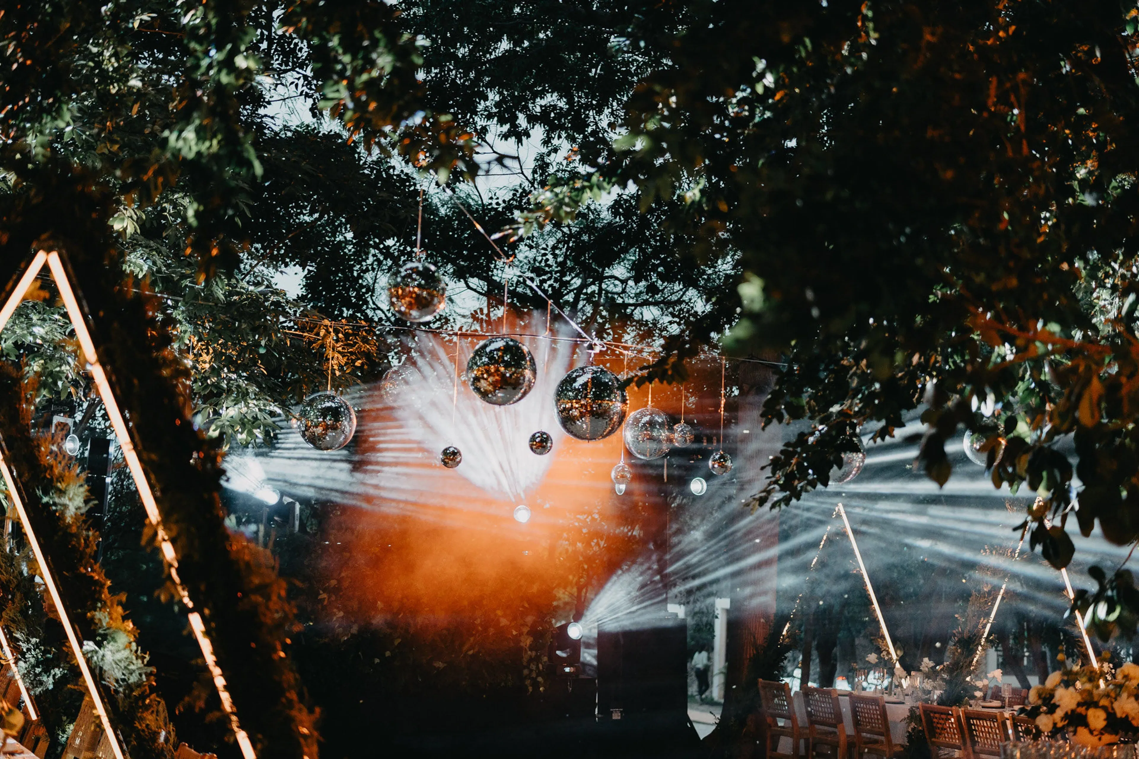 Evento al aire libre con bolas de discoteca colgando de los árboles y luces de colores que crean vibrantes patrones. Las sillas están dispuestas, lo que sugiere un ambiente festivo.