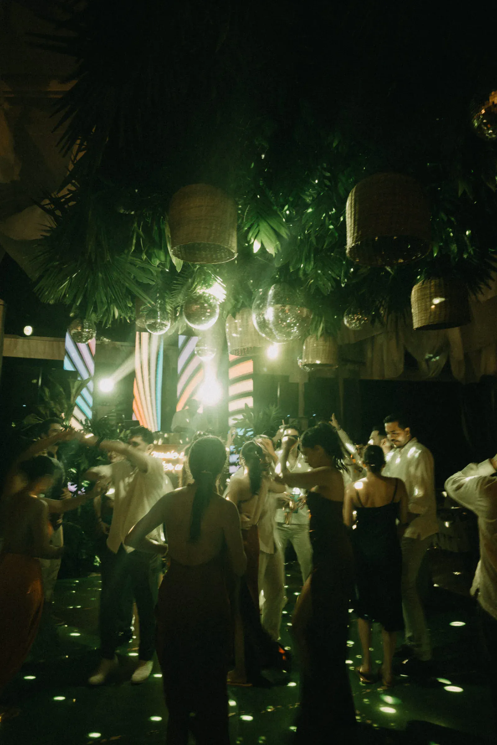Una animada fiesta con gente bailando bajo plantas colgantes y luces de mimbre. Las bolas de discoteca reflejan rayas de colores, creando un ambiente alegre.