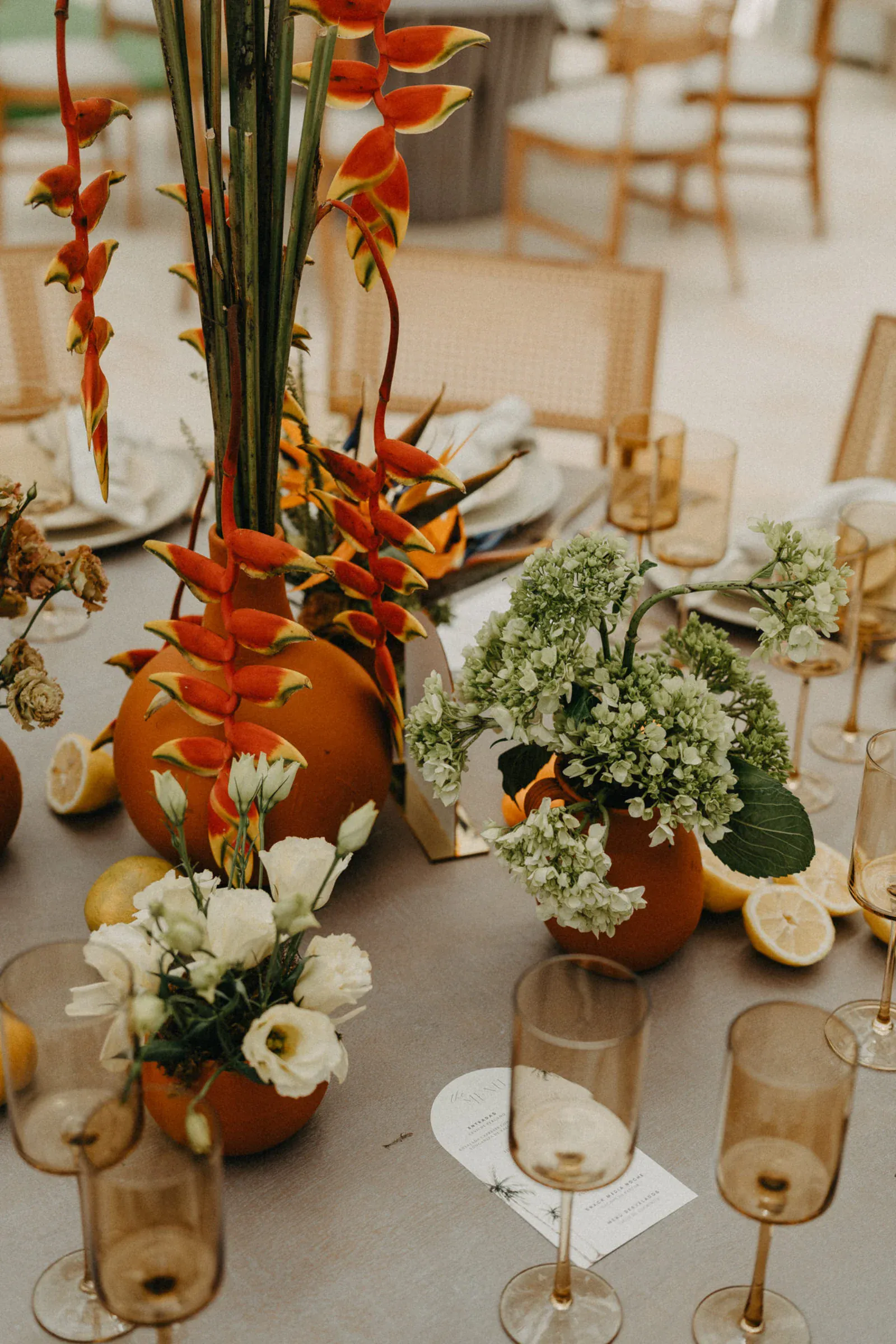 Elegante mesa con vibrantes flores tropicales rojas y amarillas en jarrones de terracota, rodeadas de flores blancas, follaje verde y limones en rodajas.