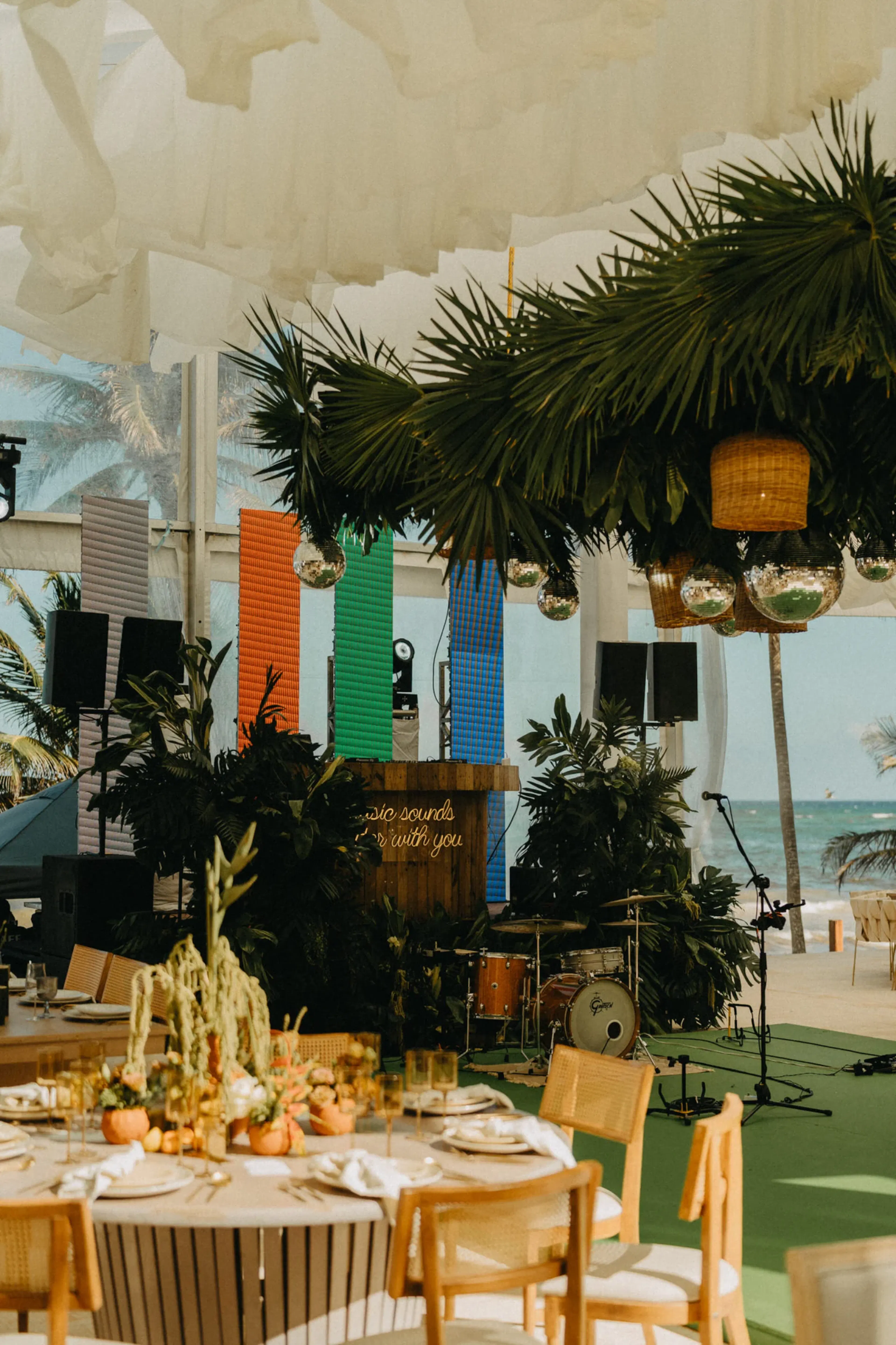 Local de temática tropical con palmeras, coloridos paneles verticales y vistas a la playa. Mesas preparadas para cenar, con un escenario y una batería cerca. Festivo y vibrante.