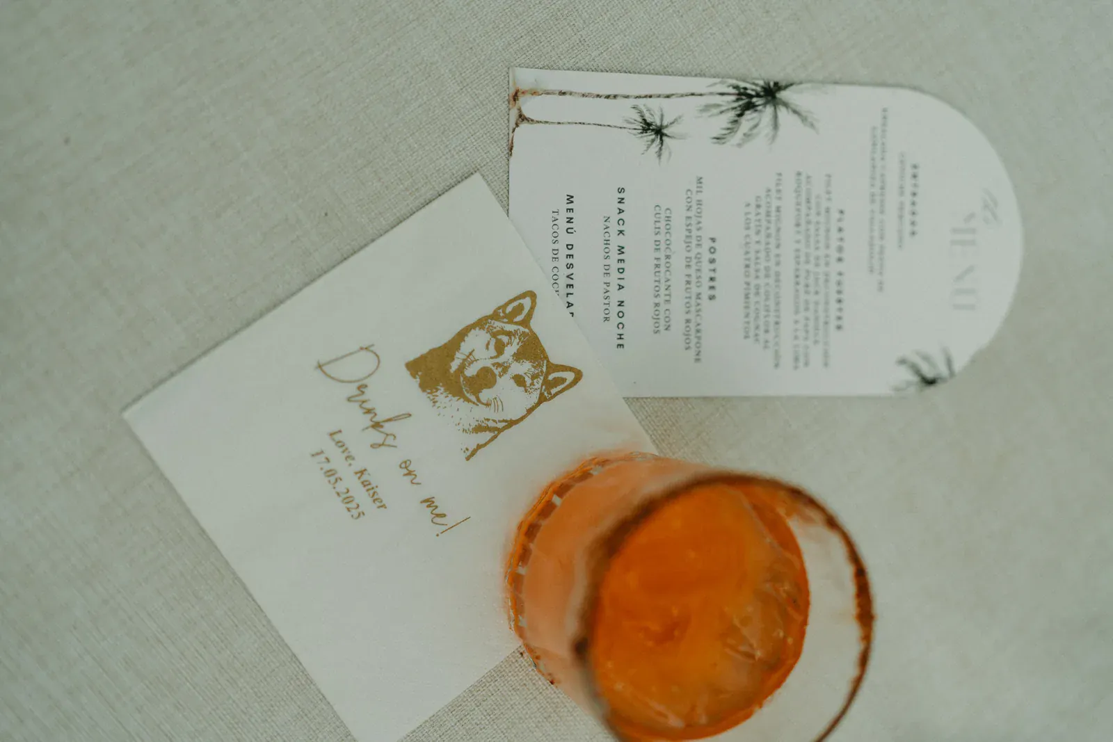Una servilleta con una ilustración de un perro y el texto "¡Las bebidas corren por mi cuenta!" junto a una tarjeta de menú blanca con palmeras y una bebida naranja sobre una superficie beige.