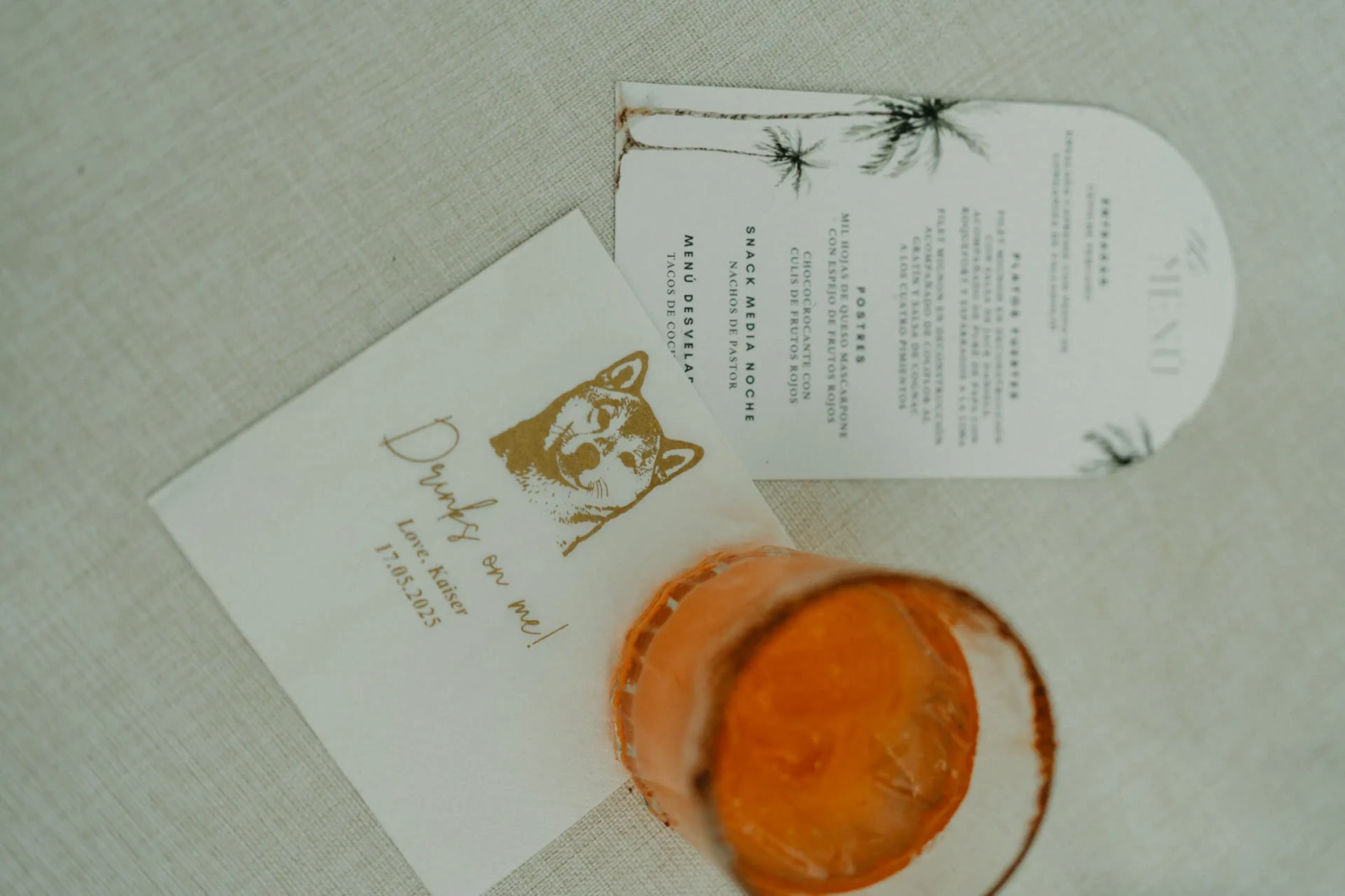 Una servilleta con una ilustración de un perro y el texto "¡Las bebidas corren por mi cuenta!" junto a una tarjeta de menú blanca con palmeras y una bebida naranja sobre una superficie beige.