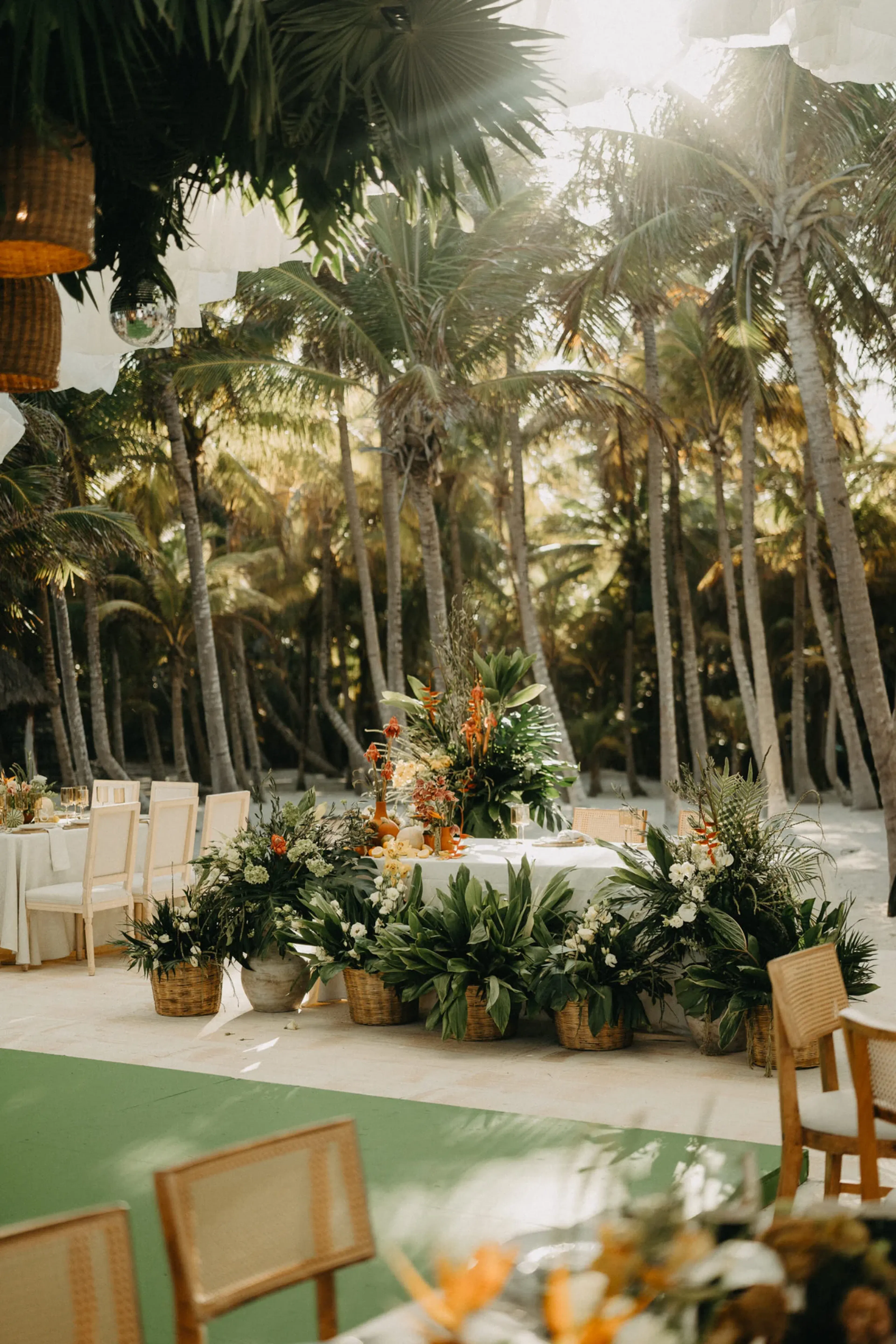 Boda tropical al aire libre bajo palmeras, con sillas y mesas adornadas con exuberante vegetación y flores vibrantes. La luz del sol se filtra entre las hojas.