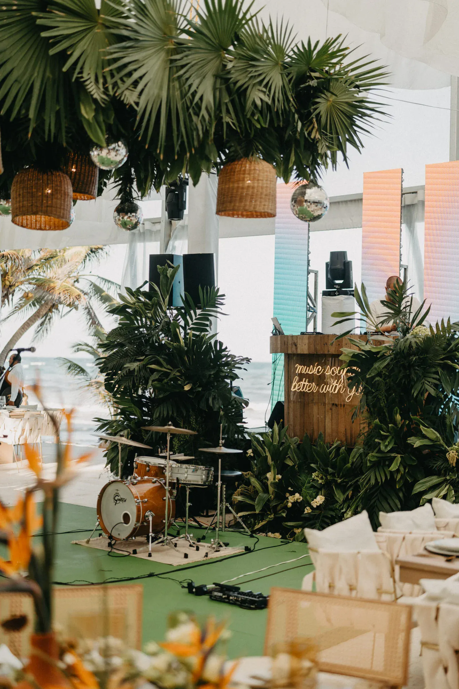 Escenario de temática tropical con tambores, exuberante vegetación, lámparas de mimbre y un letrero de neón que dice "La música suena mejor contigo". Playa y palmeras de fondo.