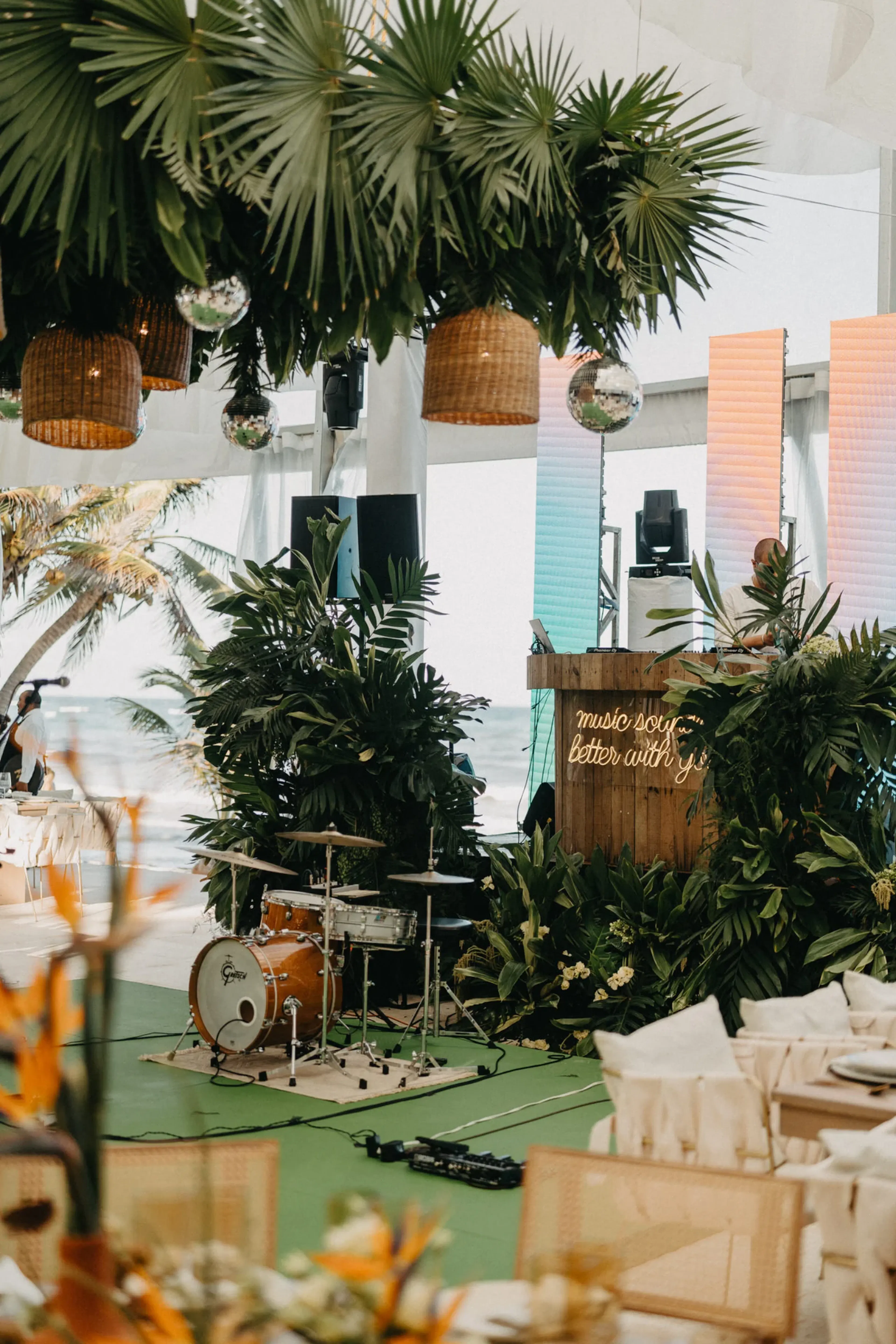 Escenario de temática tropical con tambores, exuberante vegetación, lámparas de mimbre y un letrero de neón que dice "La música suena mejor contigo". Playa y palmeras de fondo.