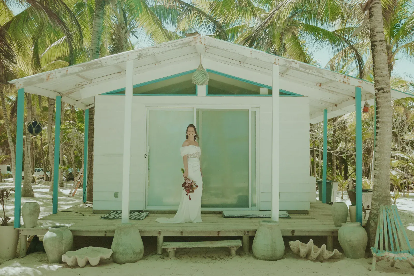 Una novia con un vestido blanco se encuentra en el porche de una pequeña cabaña en la playa, rodeada de palmeras. Sostiene un ramo de flores, evocando un ambiente sereno y tropical.