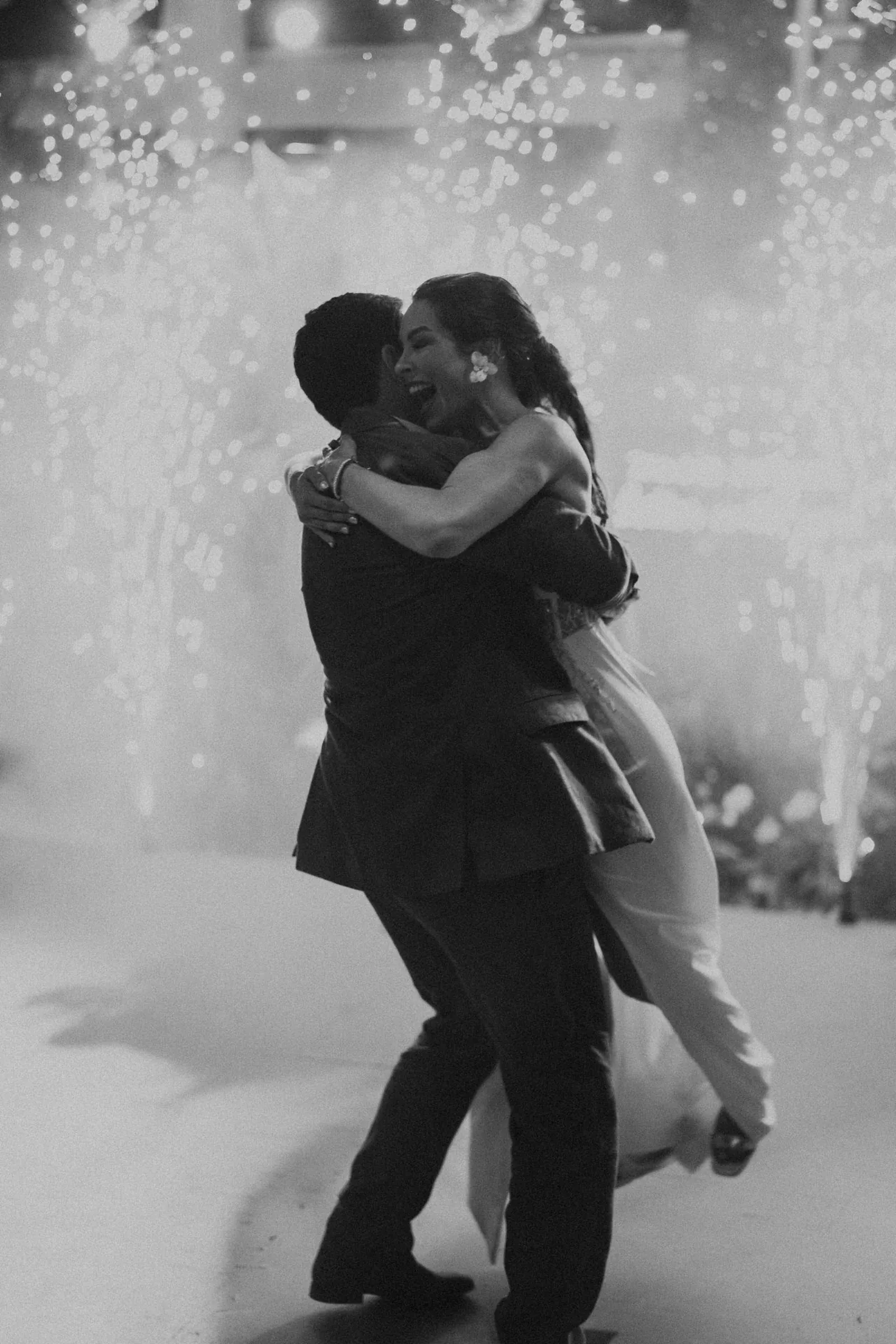 Una pareja alegre se abraza y ríe con un fondo de brillantes fuegos artificiales. La imagen en blanco y negro transmite una atmósfera festiva y romántica.