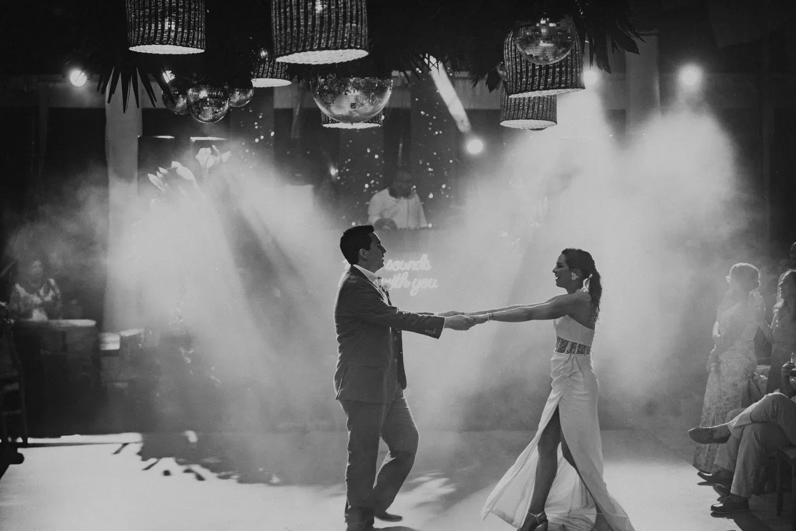 Una pareja baila con gracia en una pista llena de humo, tomados de la mano. Un DJ toca de fondo, con una iluminación suave que crea un ambiente romántico.