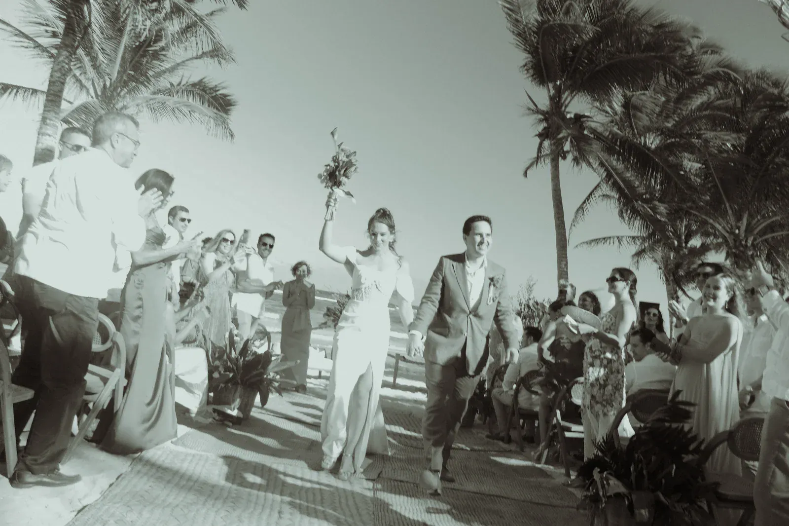 Una pareja alegre camina por un pasillo en la playa, con la novia sosteniendo un ramo en alto. Los invitados vitorean bajo las palmeras, creando un ambiente festivo.