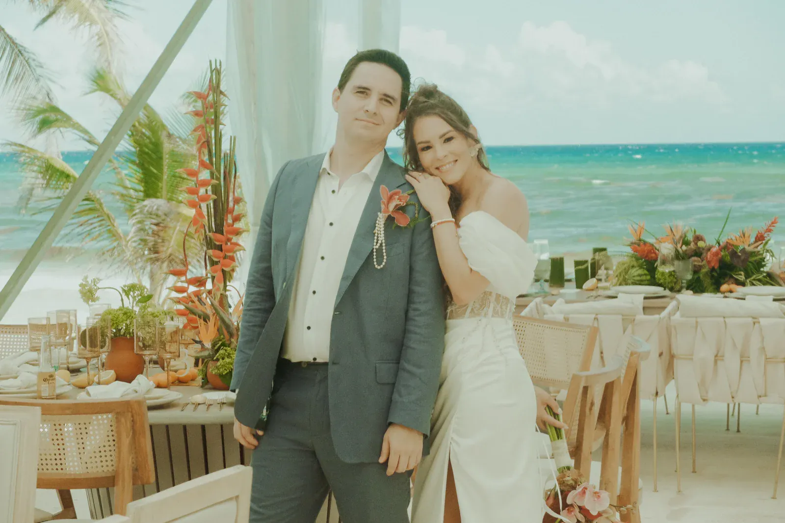 Una pareja, vestida de novia, se encuentra en una terraza junto a la playa, sonriendo cálidamente. Flores tropicales adornan las mesas, con el océano y las palmeras de fondo.