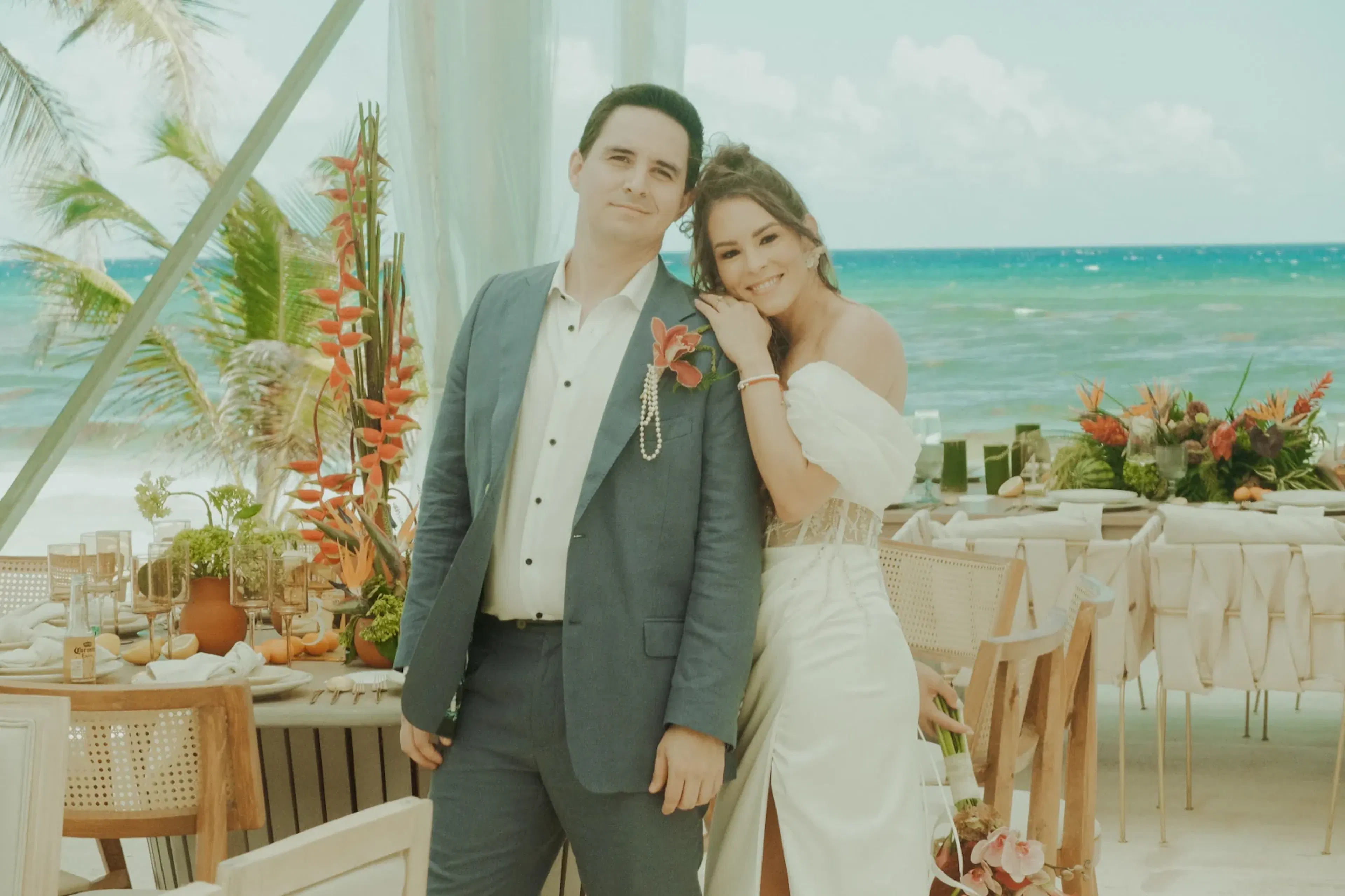 Una pareja, vestida de novia, se encuentra en una terraza junto a la playa, sonriendo cálidamente. Flores tropicales adornan las mesas, con el océano y las palmeras de fondo.
