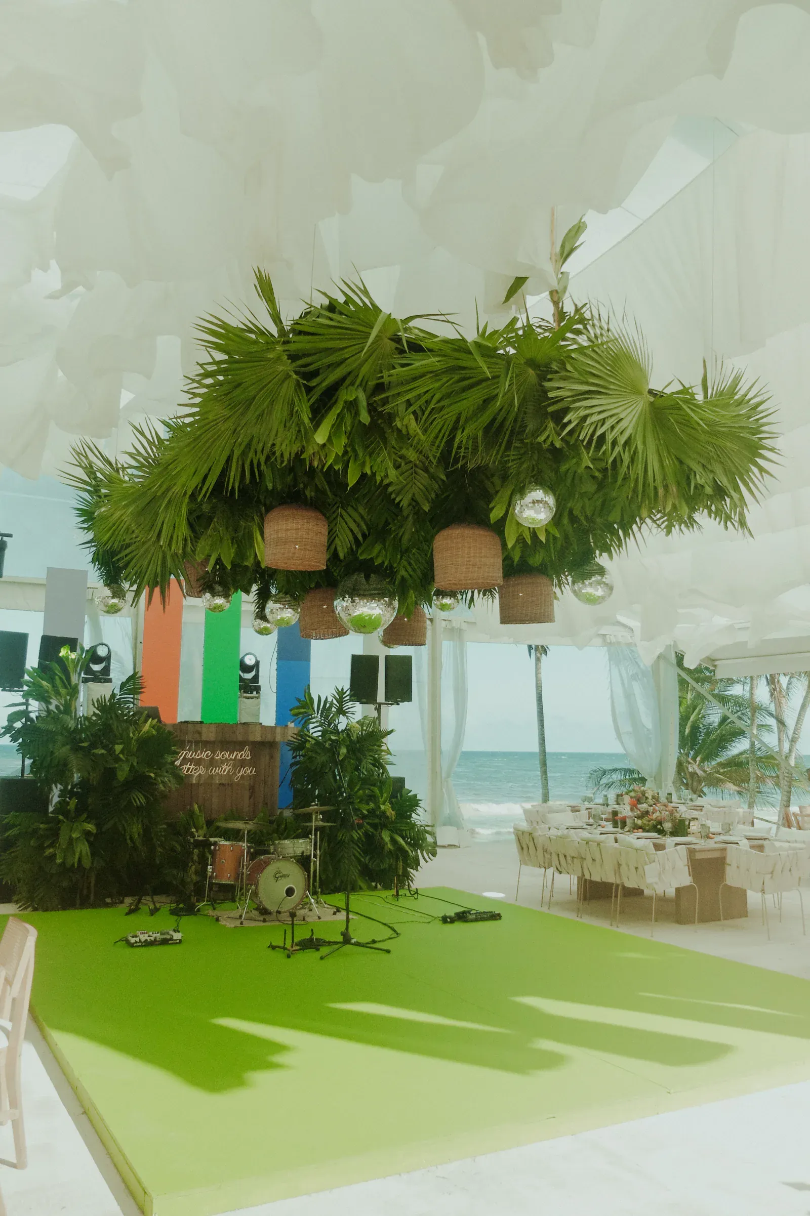 Lugar tropical para bodas junto a la playa, con exuberante vegetación y lámparas de mimbre. Cortinas blancas ondean sobre el escenario, vista al mar de fondo y mesas preparadas para cenar.