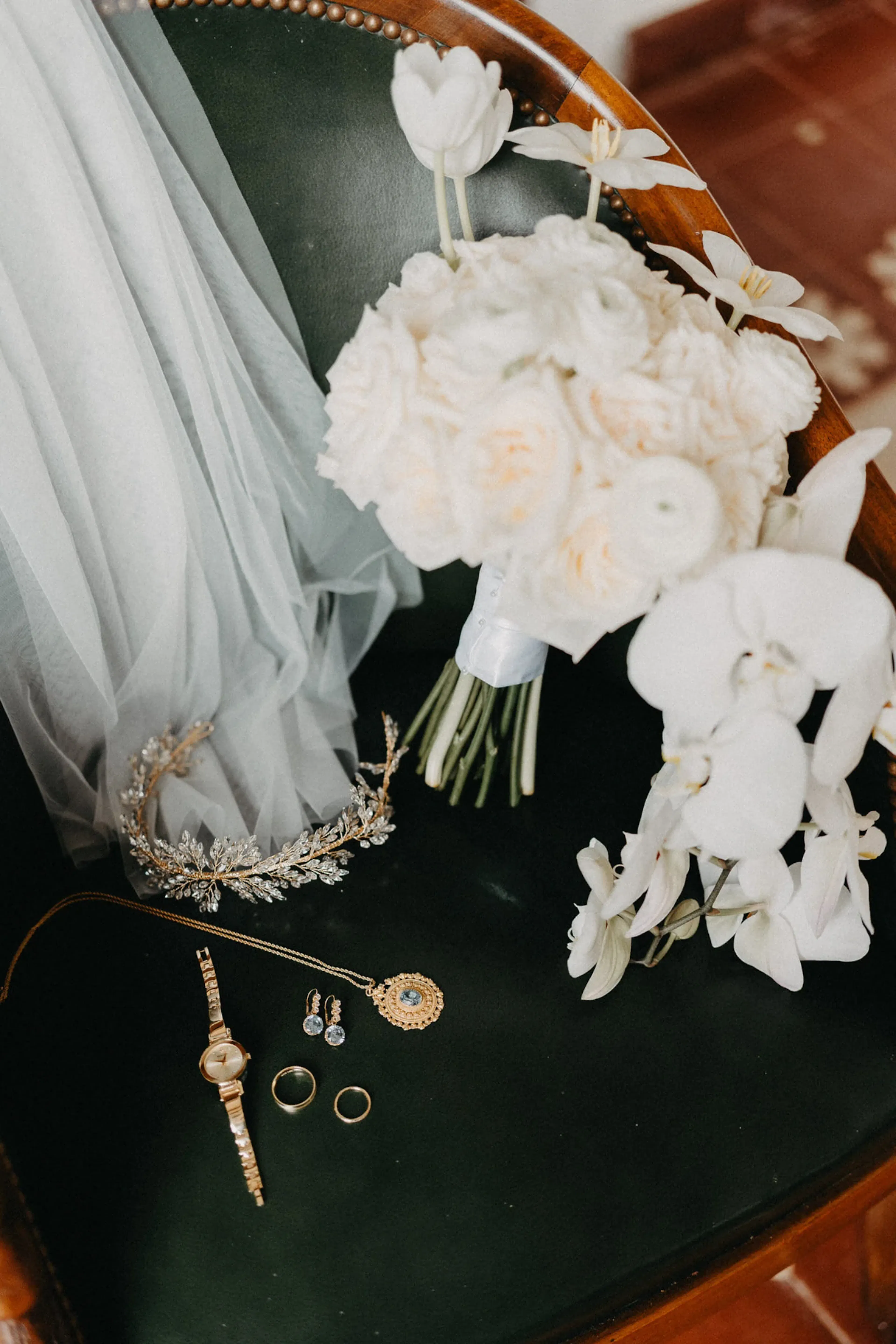 Ramo de flores blancas y velo sobre una silla verde, junto a elegantes joyas de novia que incluyen un collar, pendientes, anillos y un reloj, transmitiendo la elegancia de una boda.
