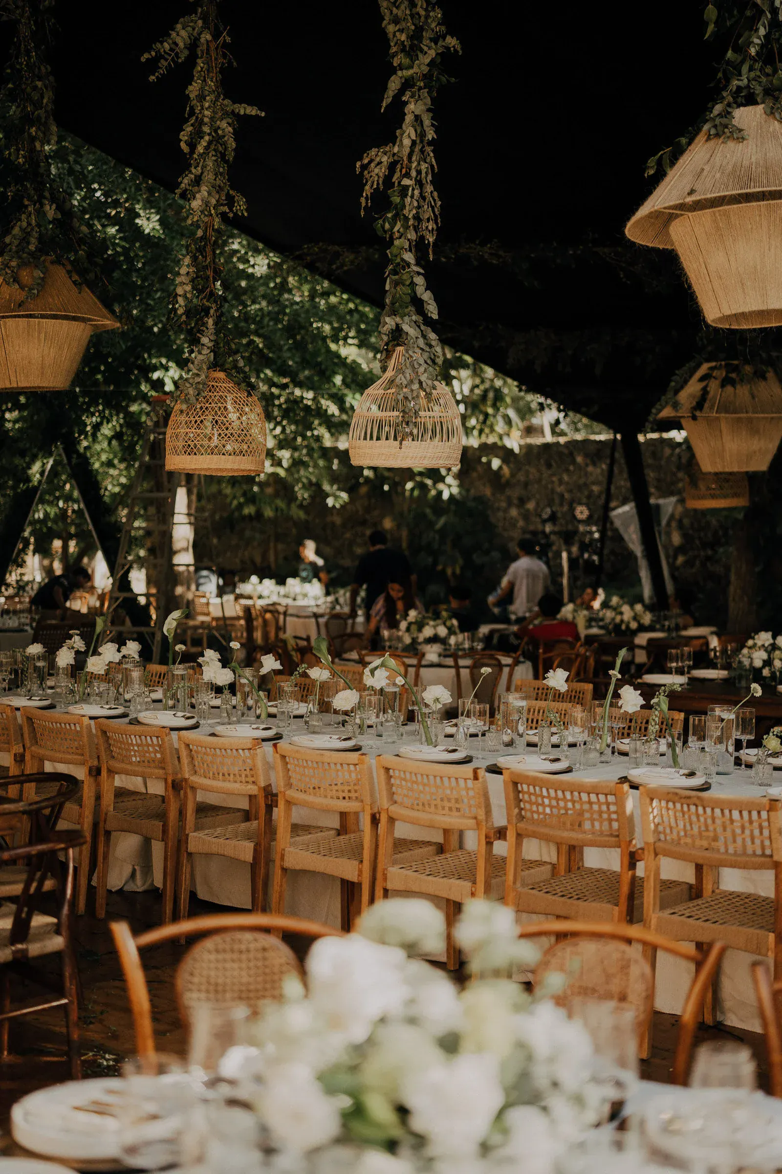 Escena de boda rústica al aire libre con largas mesas cubiertas de blanco, sillas de madera y lámparas de mimbre colgantes. La vegetación realza el ambiente cálido y acogedor.